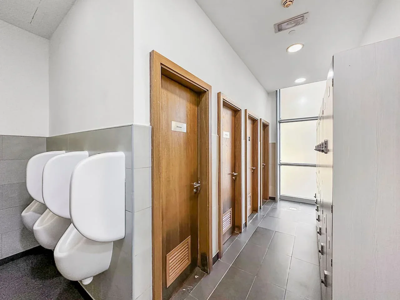 Un pasillo de baño alicatado con urinarios junto a la pared y puertas de madera.