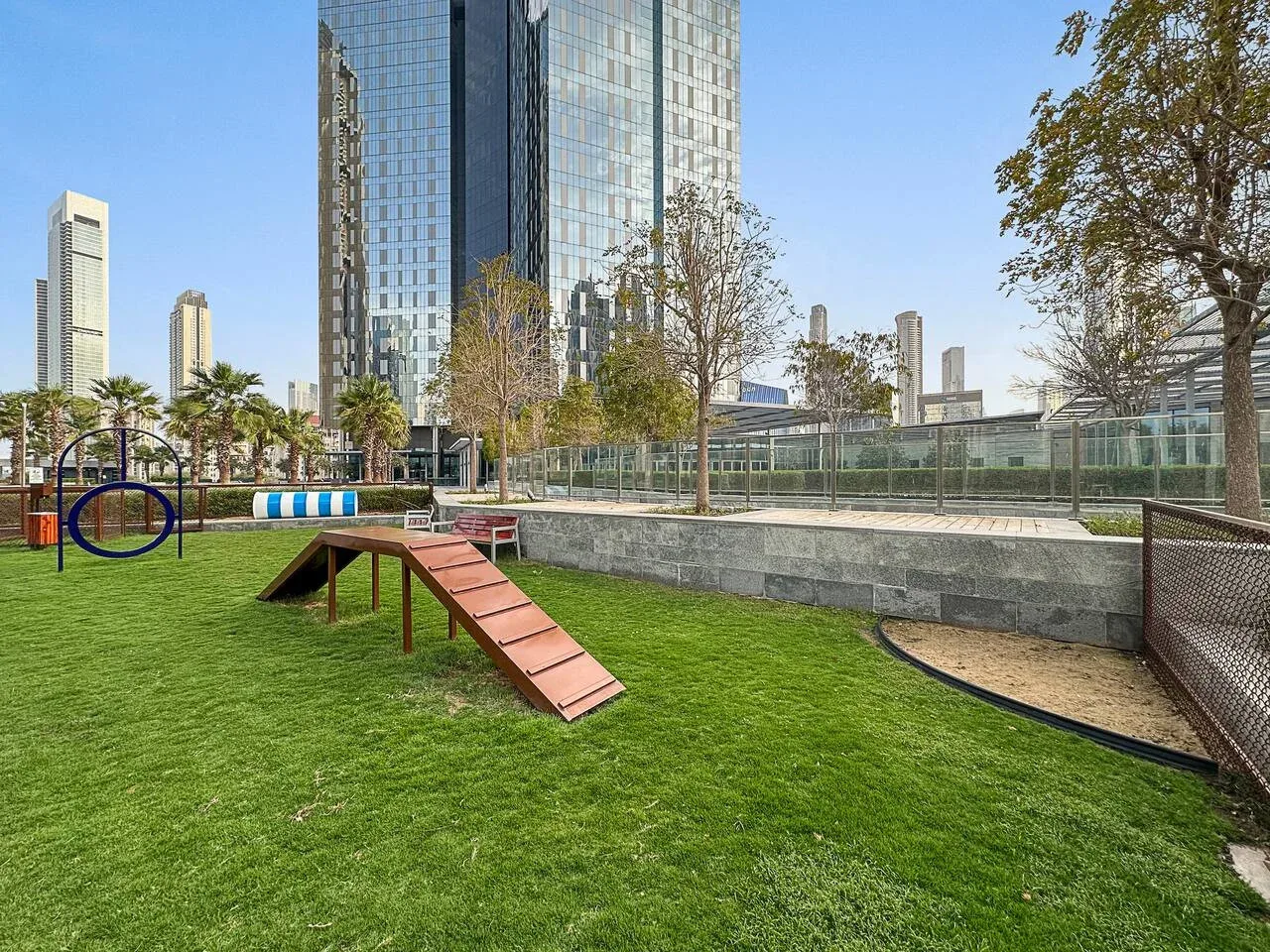 Una zona de juegos al aire libre tiene césped, una pequeña rampa de madera y un arenero cercado.