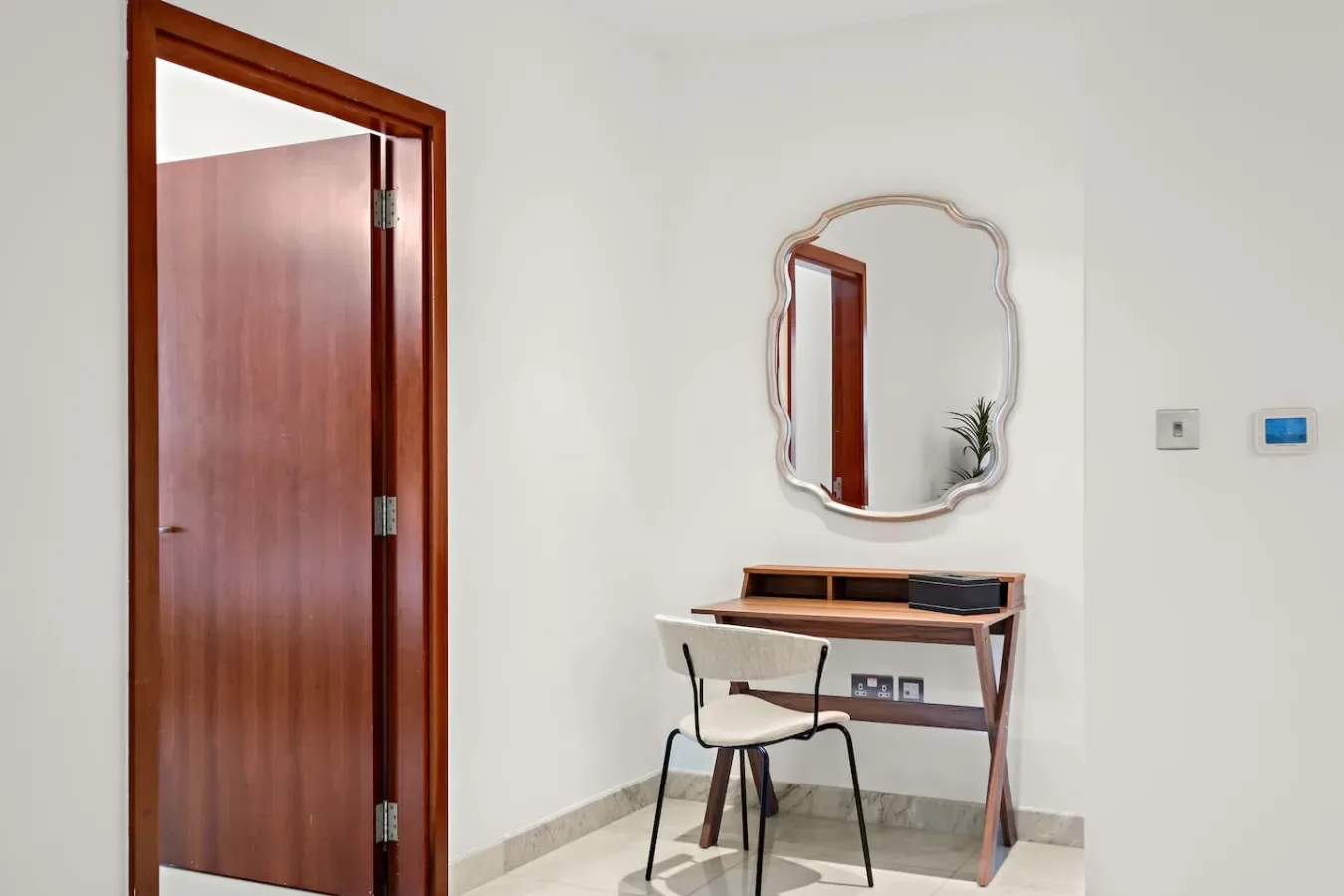 Un escritorio pequeño y una silla están bajo un espejo decorativo en una habitación luminosa.