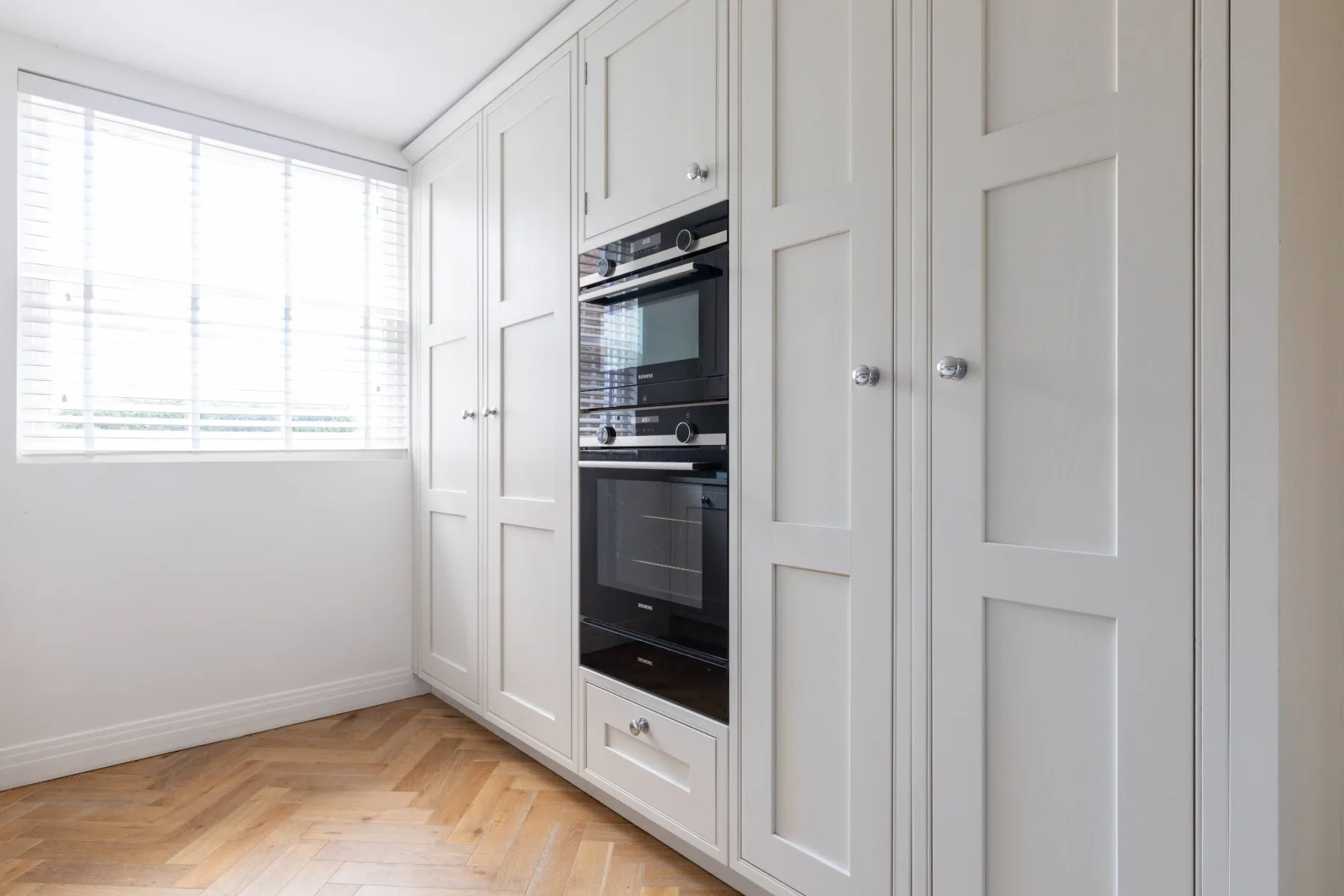 A bright kitchen with white cabinets and built-in ovens beside a large window.