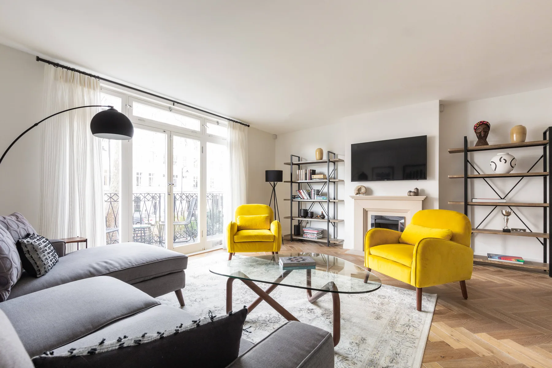 A bright living room with yellow seating, a glass coffee table, and tall windows.