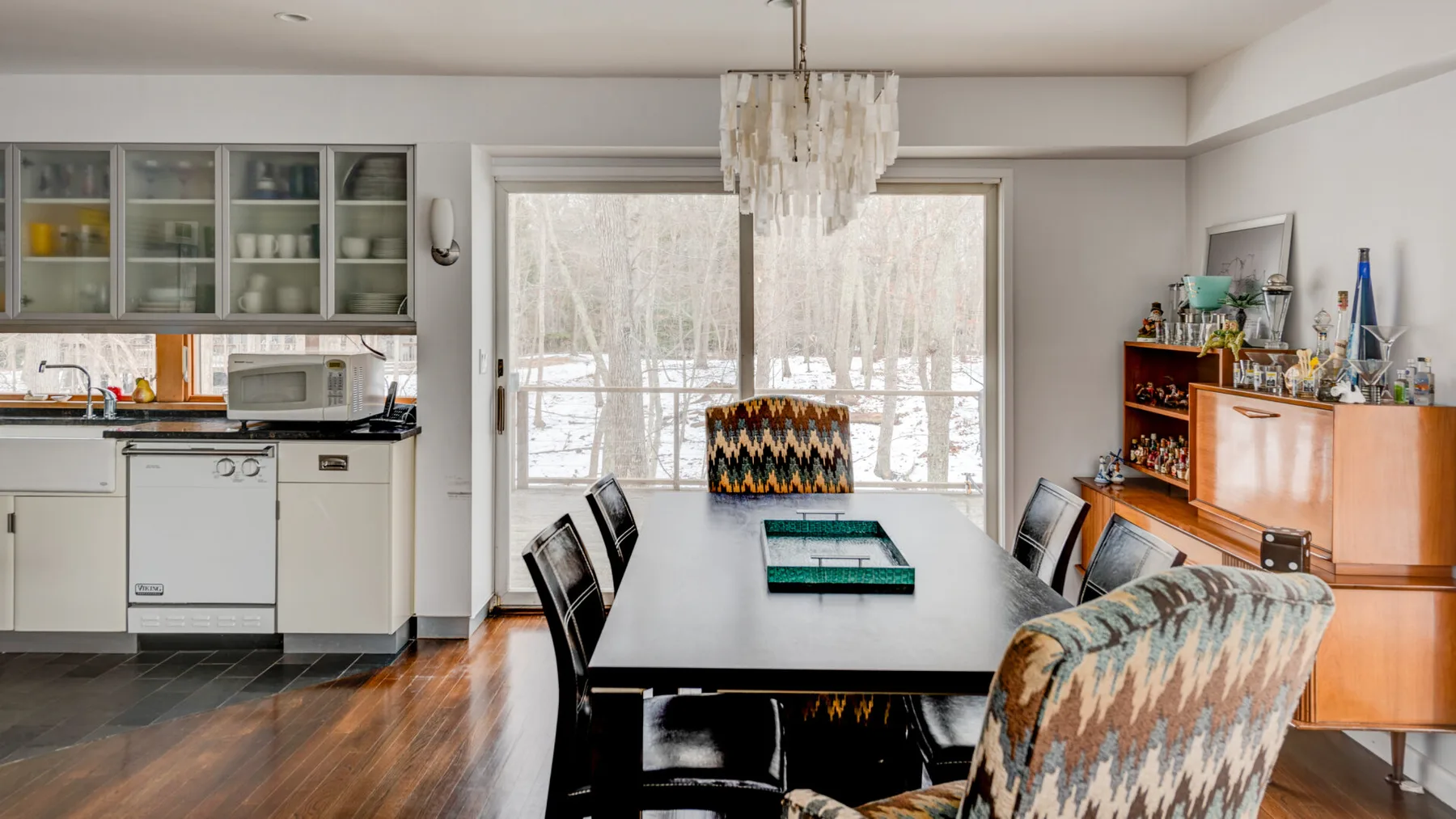 A dining area with a long table beside a kitchen and large windows.