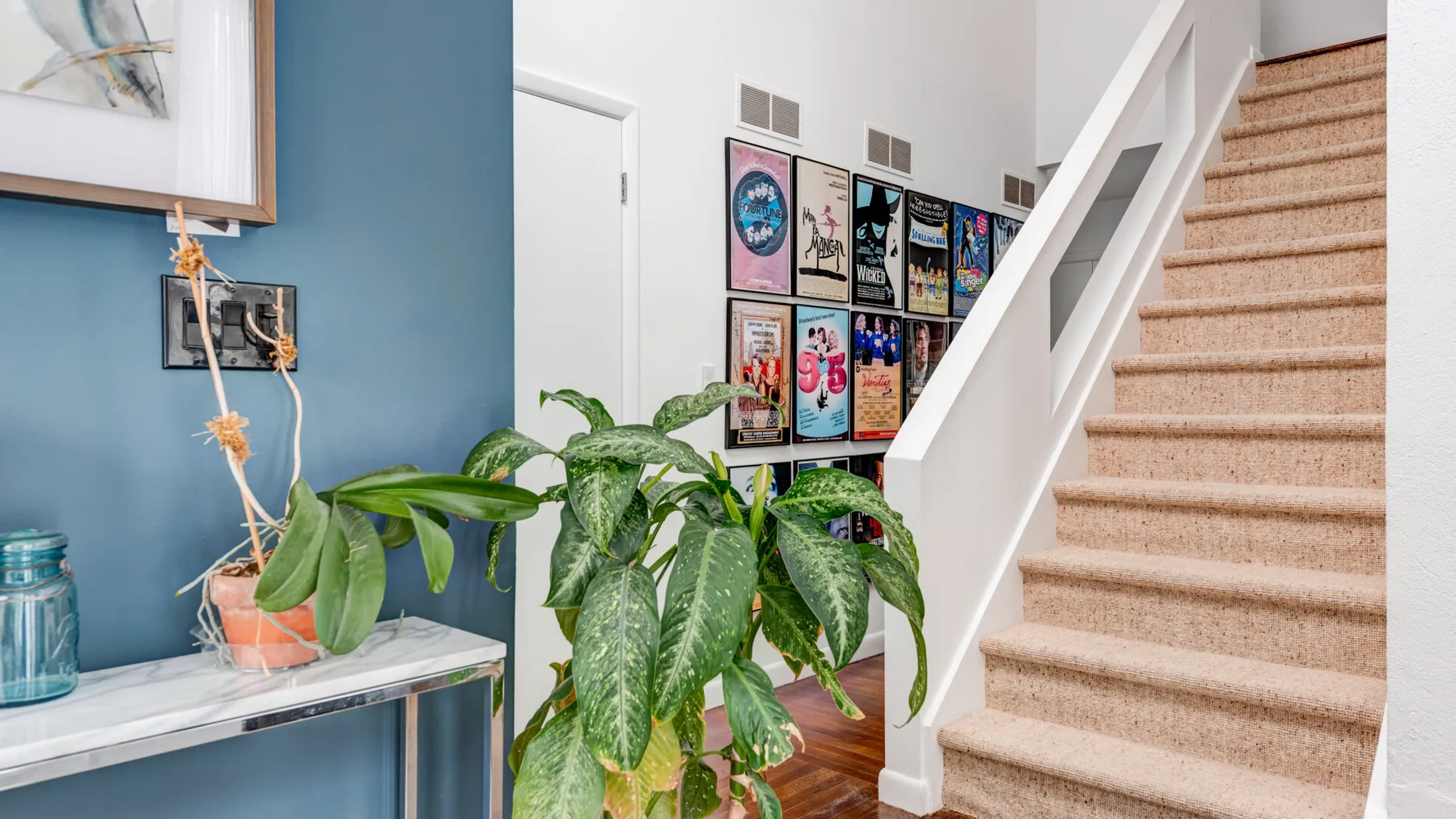 A carpeted staircase rises beside a blue wall decorated with framed posters and plants.
