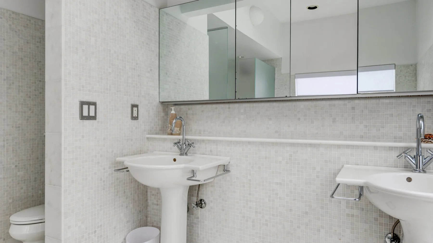 A bright tiled bathroom with pedestal sinks and a wide wall mirror.
