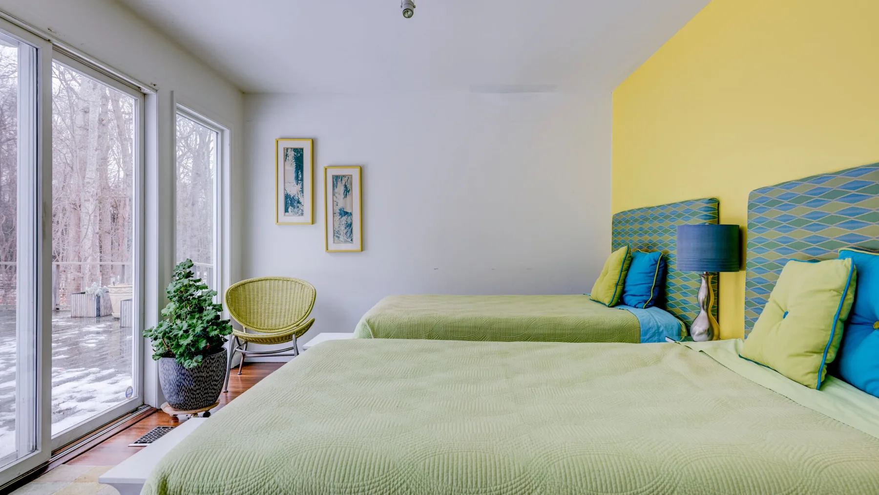A bright bedroom with two beds, colorful cushions, and tall windows.