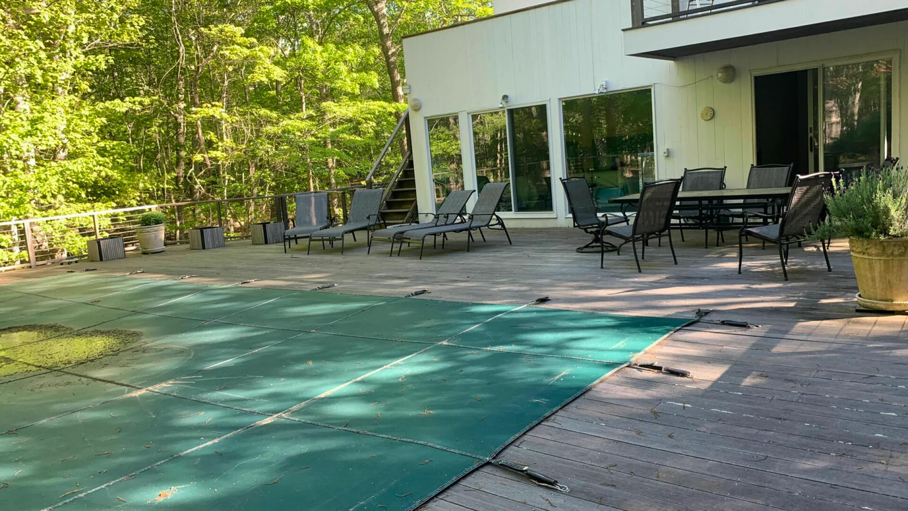 An outdoor deck beside a swimming pool, with patio seating and a wooded backdrop.