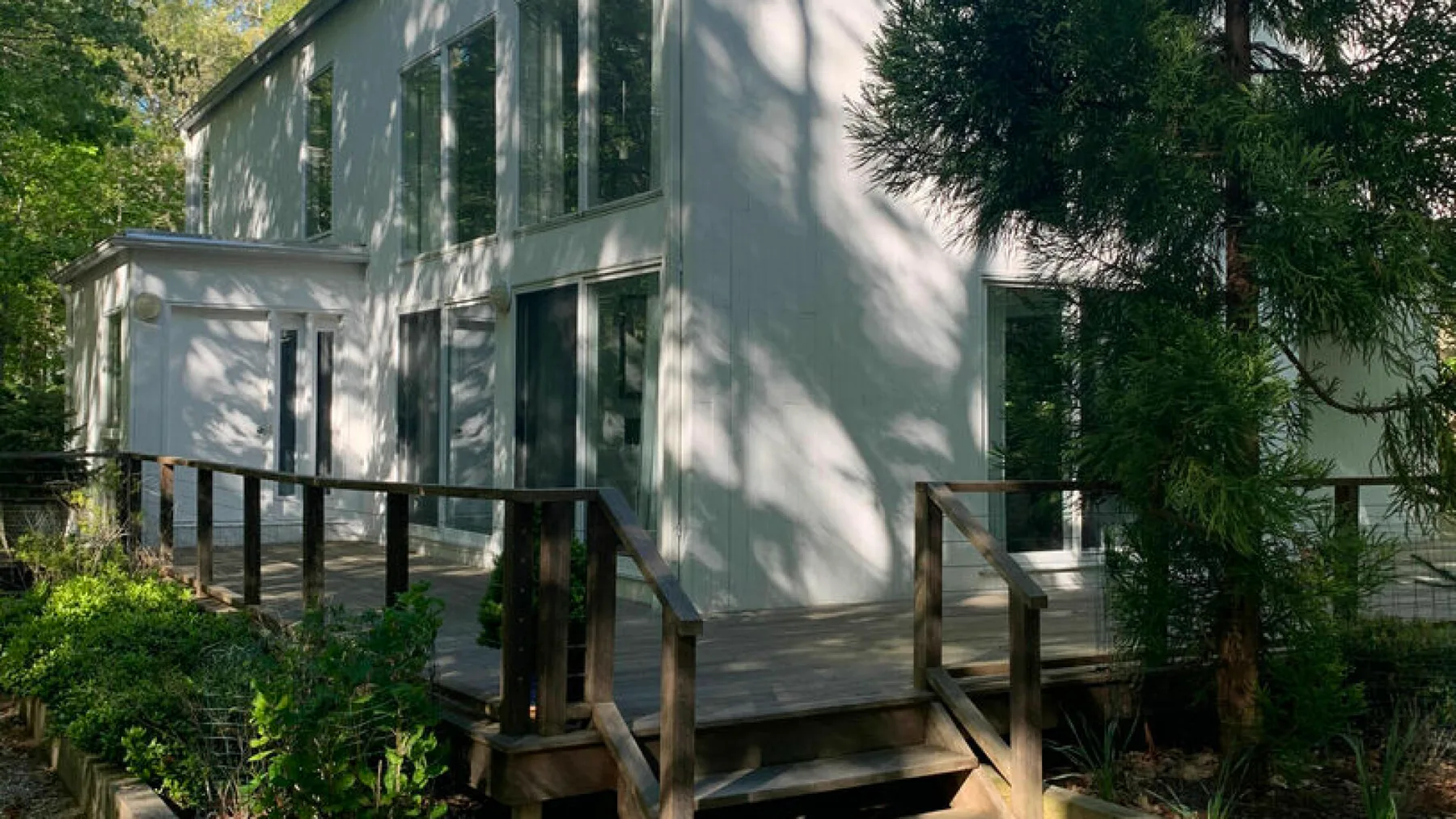 A modern white house with large windows and a wooden deck surrounded by trees.