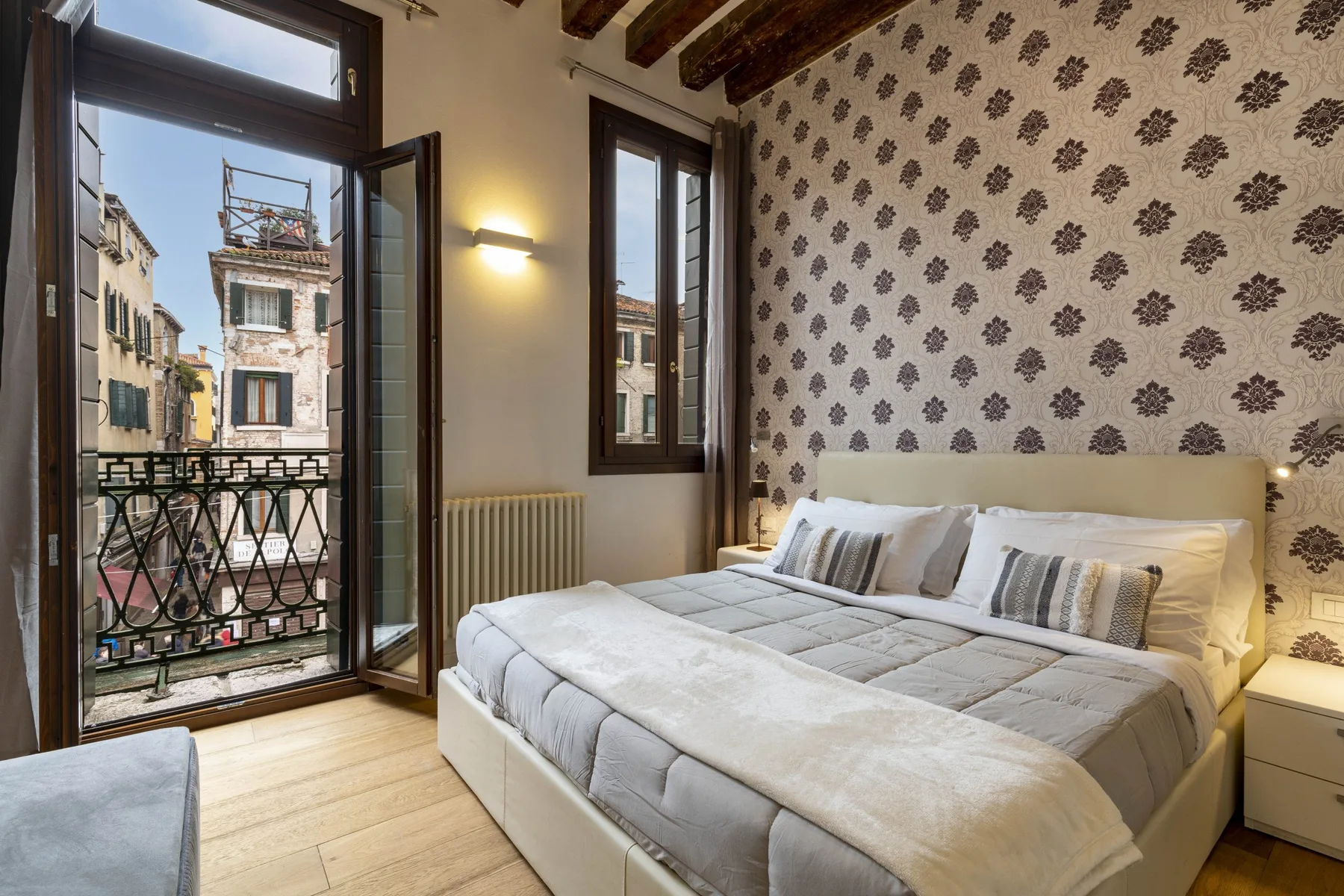 A bedroom with a double bed, patterned wallpaper, and tall windows overlooking neighboring buildings.