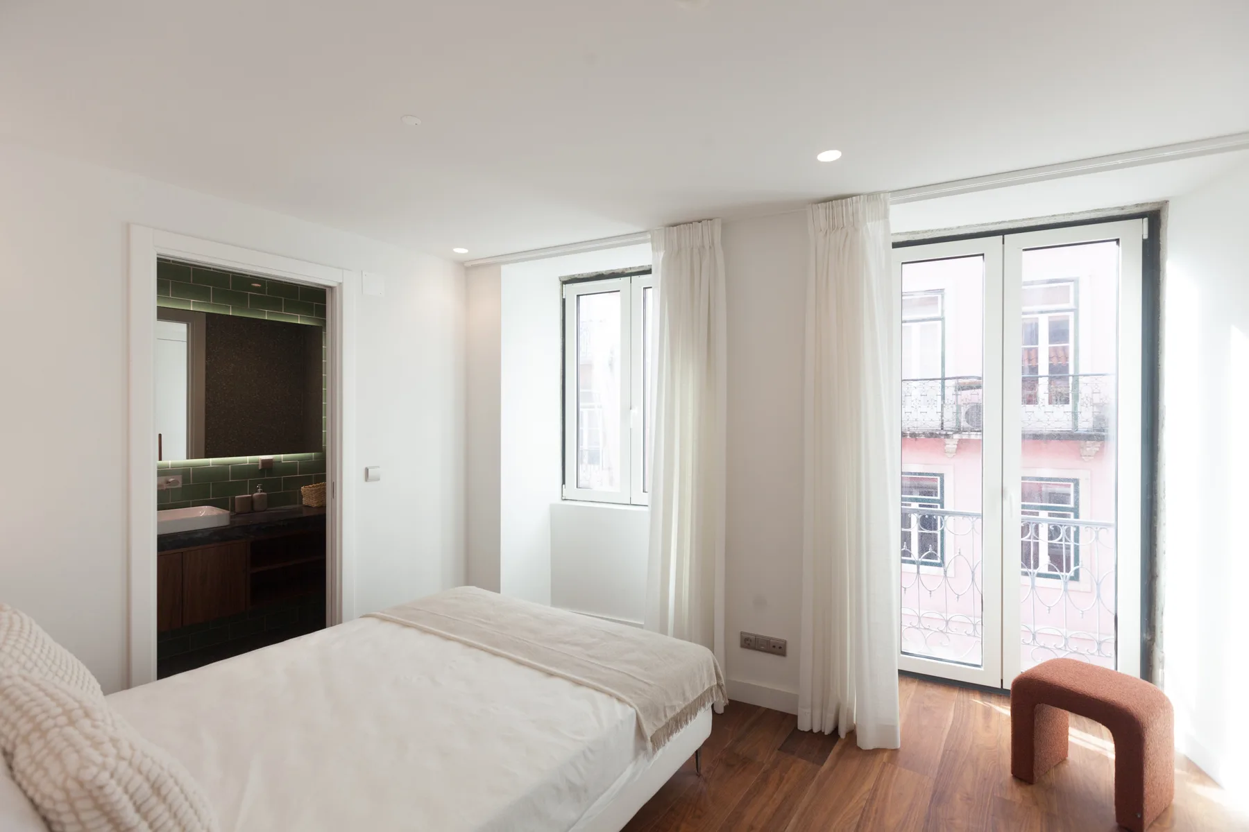 Un dormitorio luminoso con una cama junto a ventanas altas y cortinas claras.