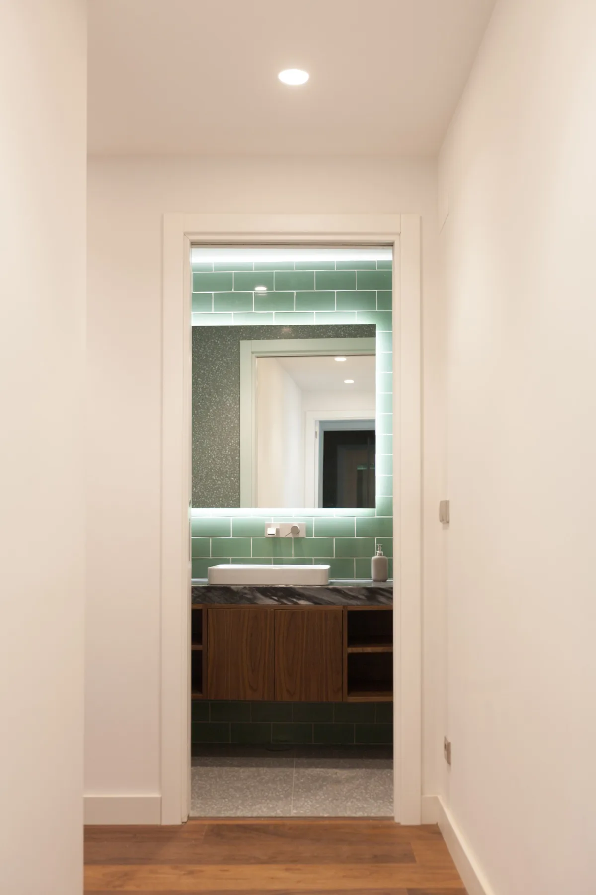 Un pasillo da a un baño con una pared de azulejos verdes y un lavabo.