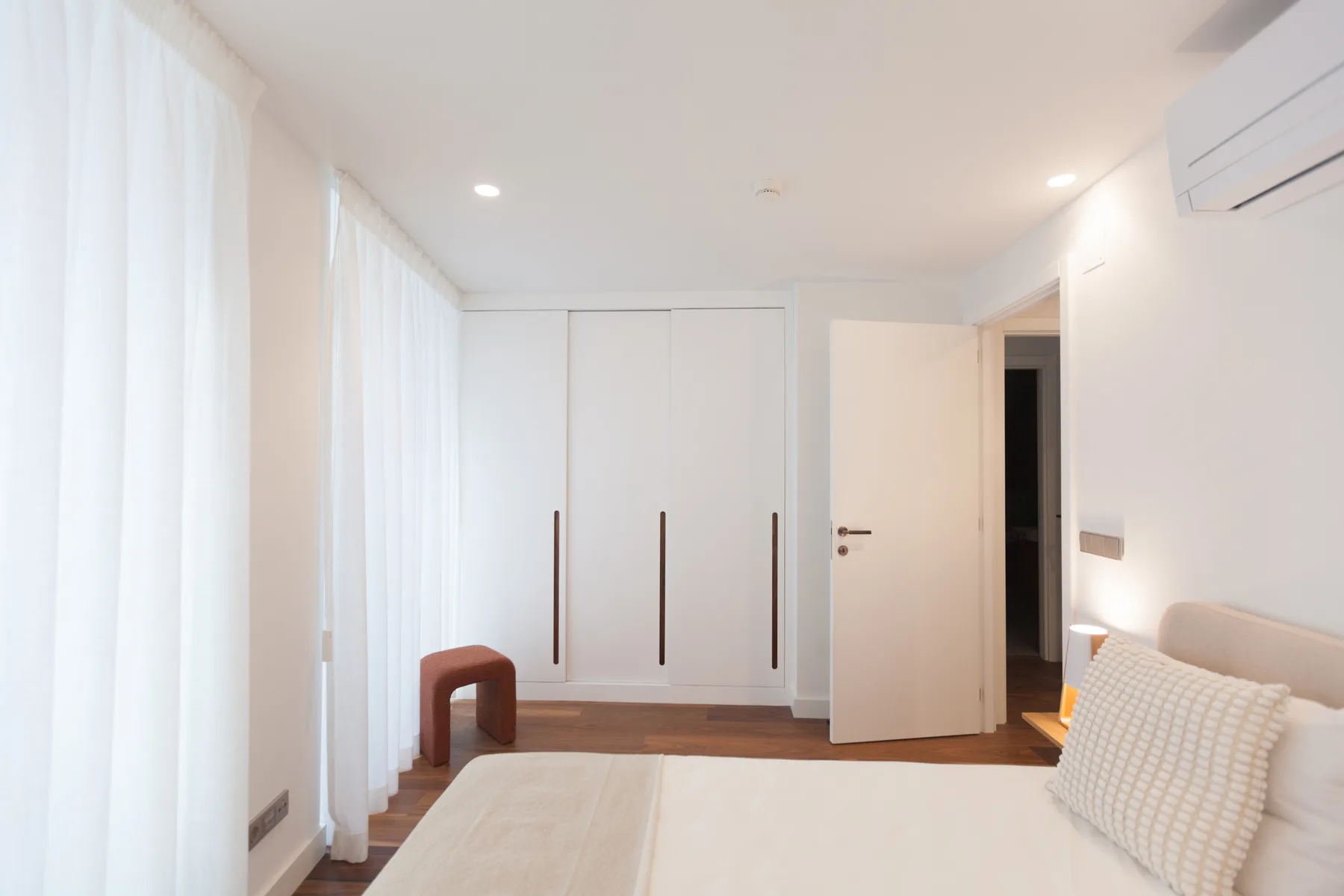 Un dormitorio luminoso con una cama, cortinas translúcidas y un armario blanco.
