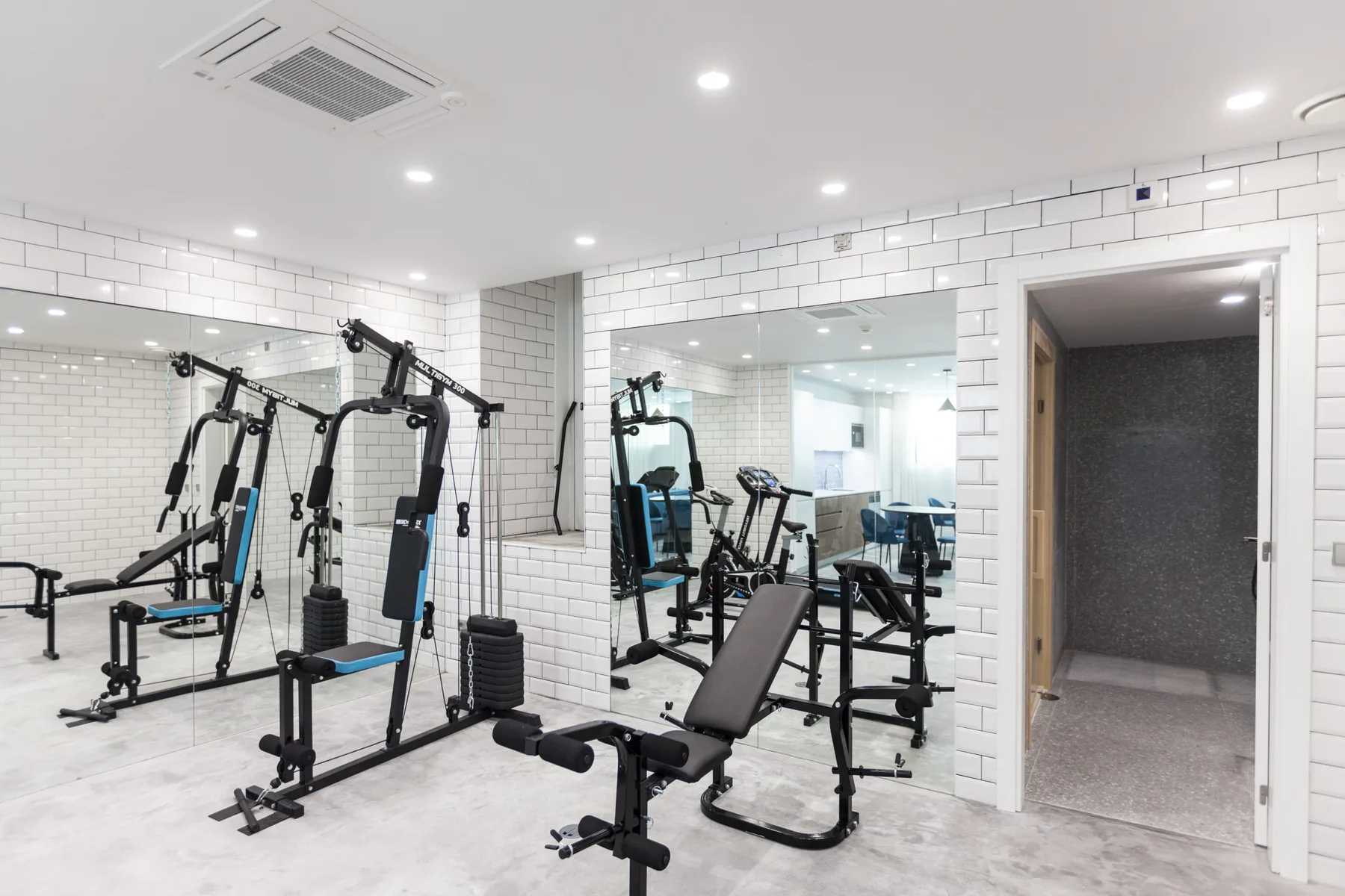 Sala de ejercicio con azulejos, máquinas de pesas, bancos y una pared de espejos.