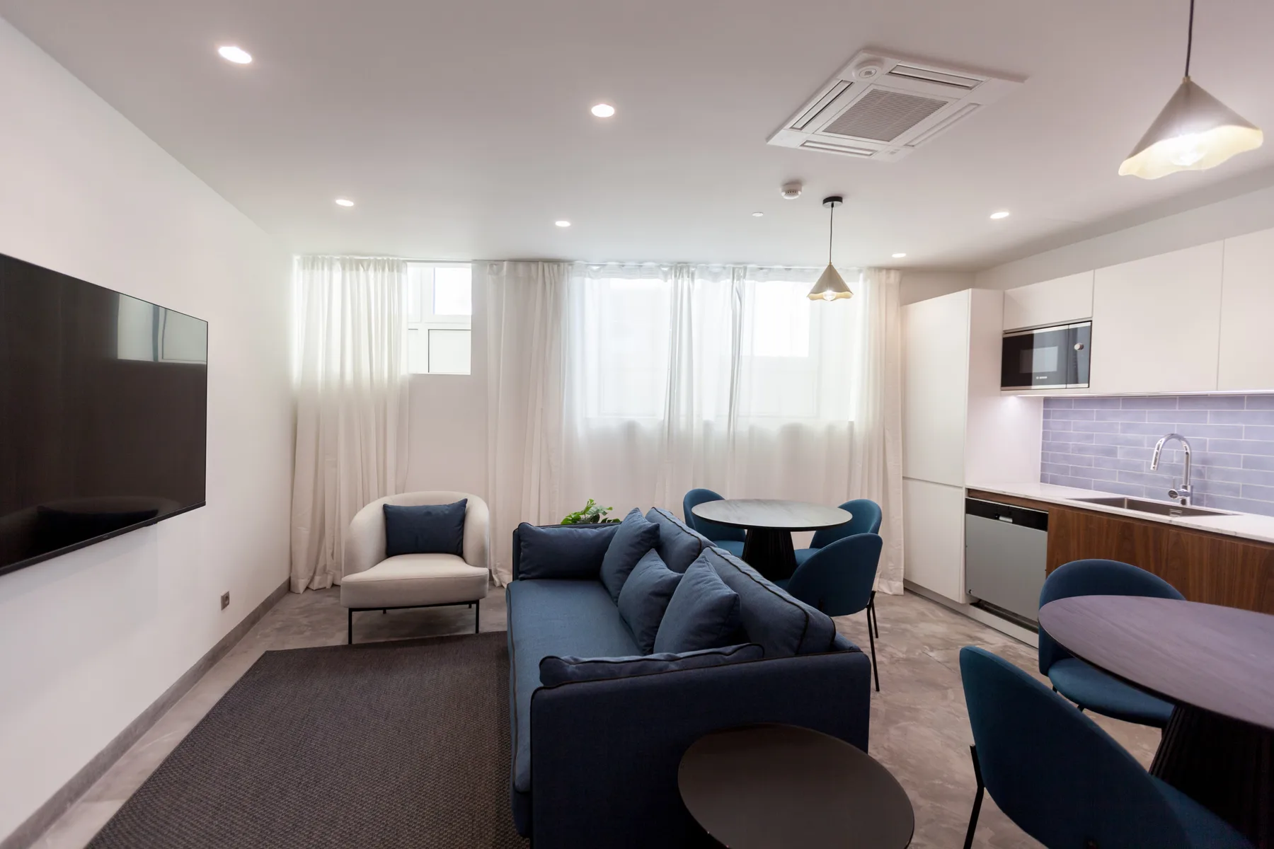 Una luminosa sala de estar y comedor tiene asientos azules, un televisor y una cocina compacta.
