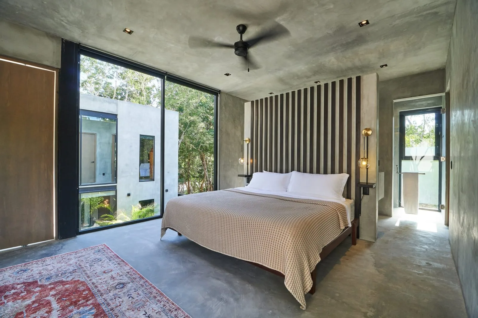 Un dormitorio con una cama bien tendida y grandes ventanas con vistas a la vegetación.
