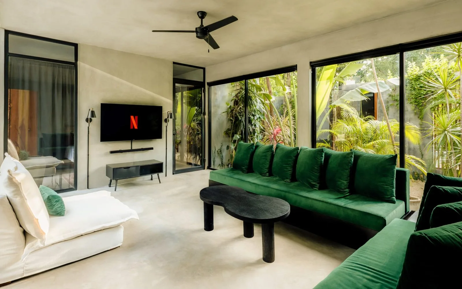 A bright living room with green seating and floor-to-ceiling windows overlooking lush plants.