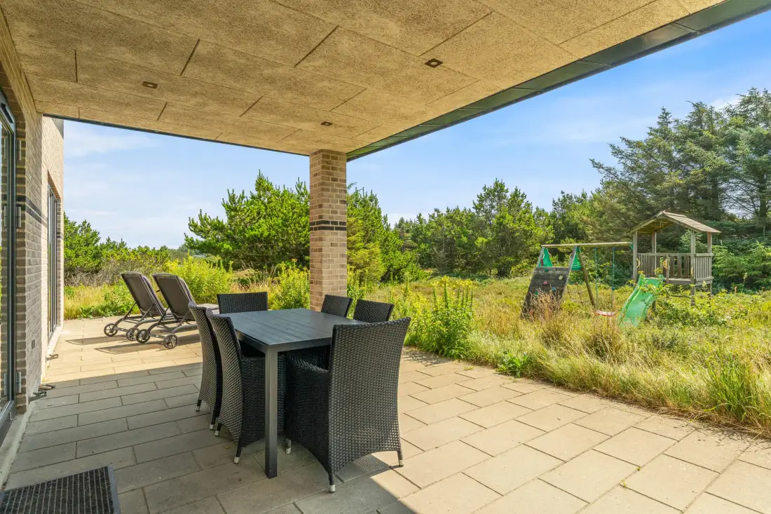 Un patio cubierto tiene una mesa de comedor y asientos junto a un jardín con césped.