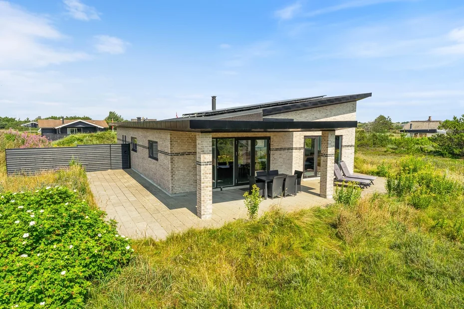 Una casa moderna con patio pavimentado y asientos al aire libre está rodeada de vegetación.