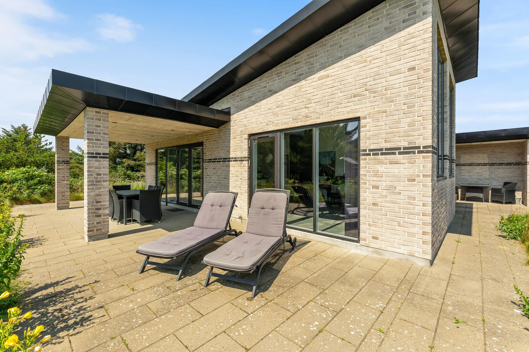 Un patio junto a una casa moderna tiene asientos reclinables y una zona de comedor cubierta.
