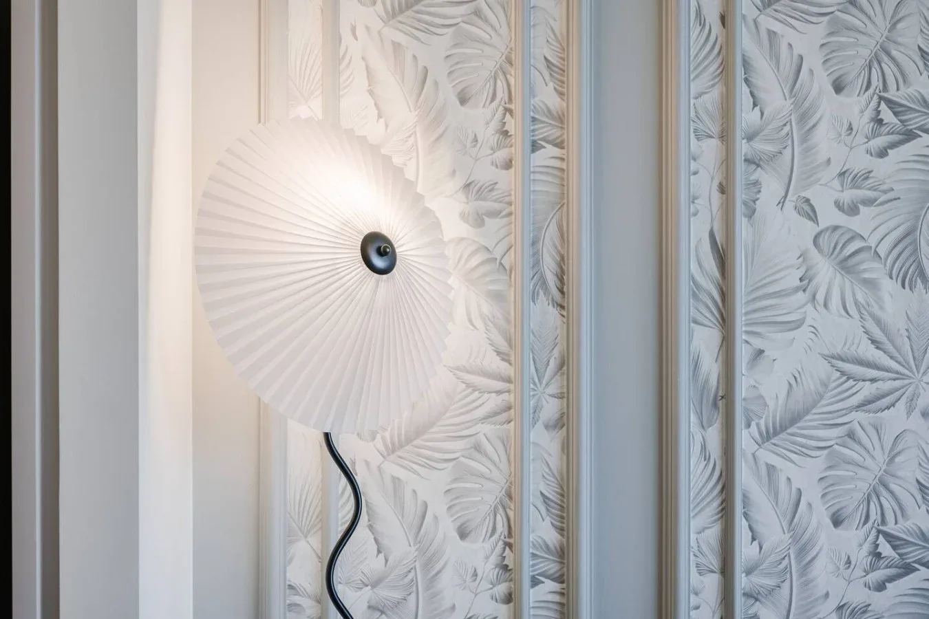 Una lámpara de pared está frente a un papel tapiz claro con motivos botánicos.