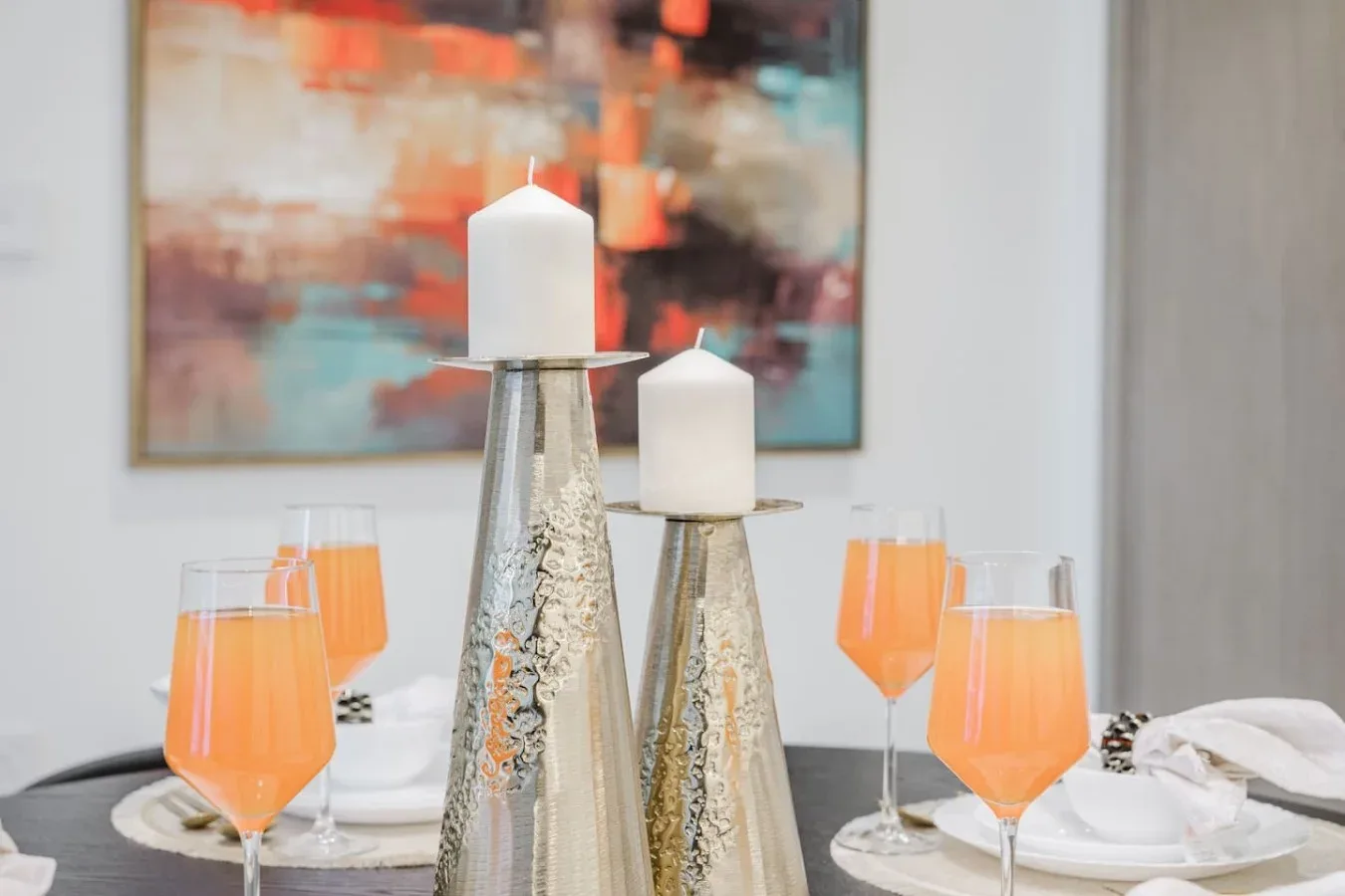 Una mesa de comedor está preparada con bebidas naranjas y velas decorativas bajo un cuadro colorido.