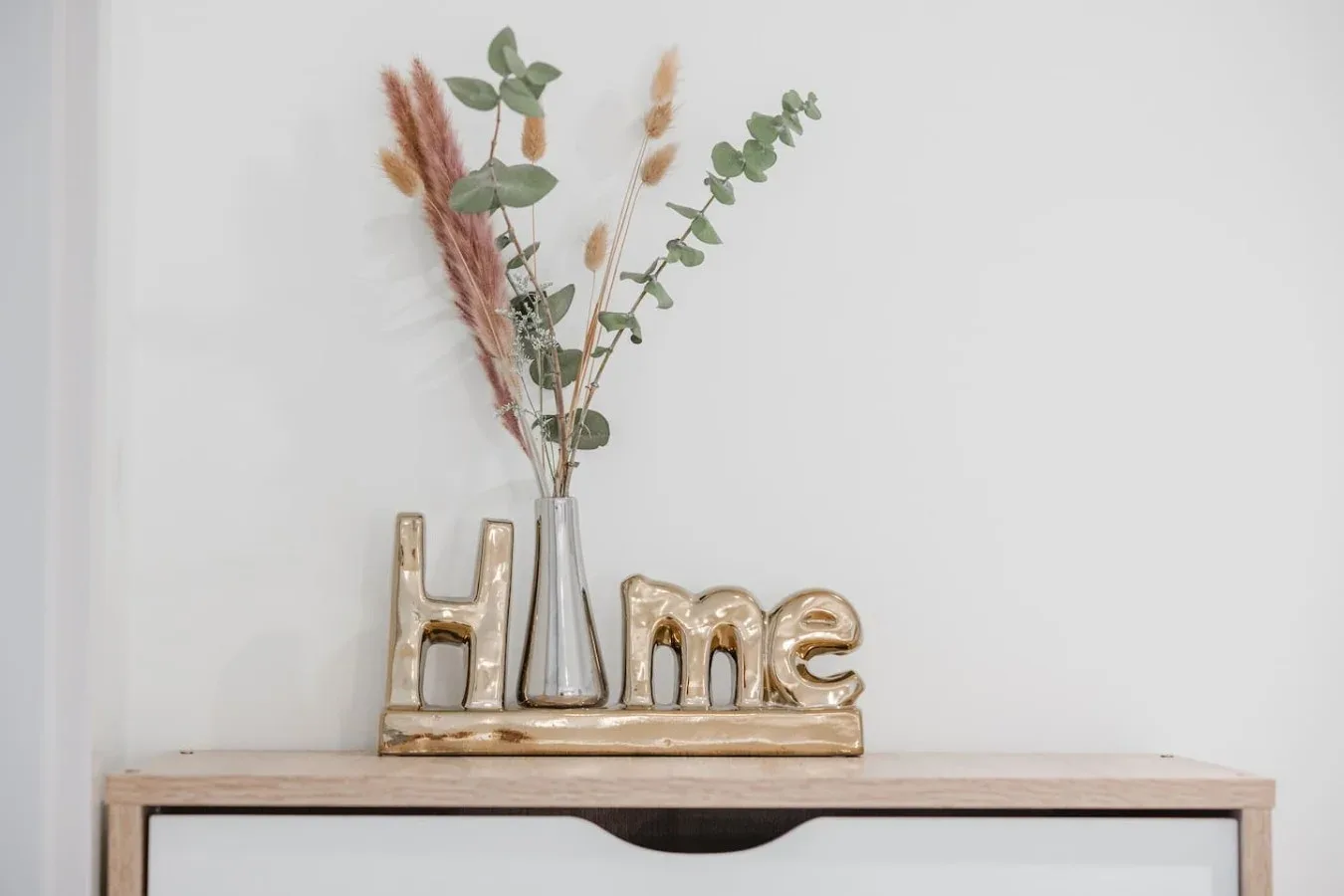 Un jarrón decorativo y un adorno dorado con la palabra «Home» están sobre un estante estrecho, junto a una pared blanca.