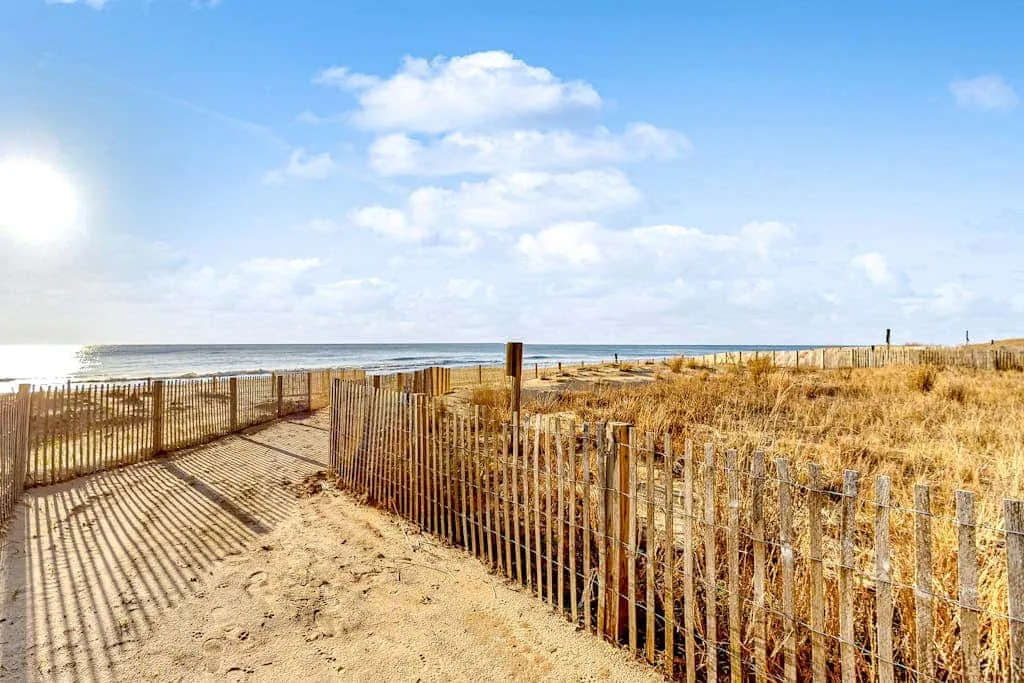 A sandy path runs between dune grasses and wooden fencing toward the ocean.