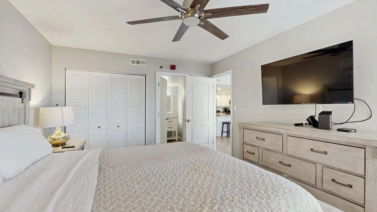 A bright bedroom with a neatly made bed, dresser, and wall-mounted television.
