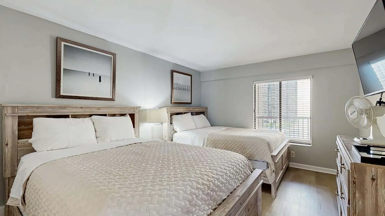 A bedroom with two beds, light bedding, and a window with blinds.