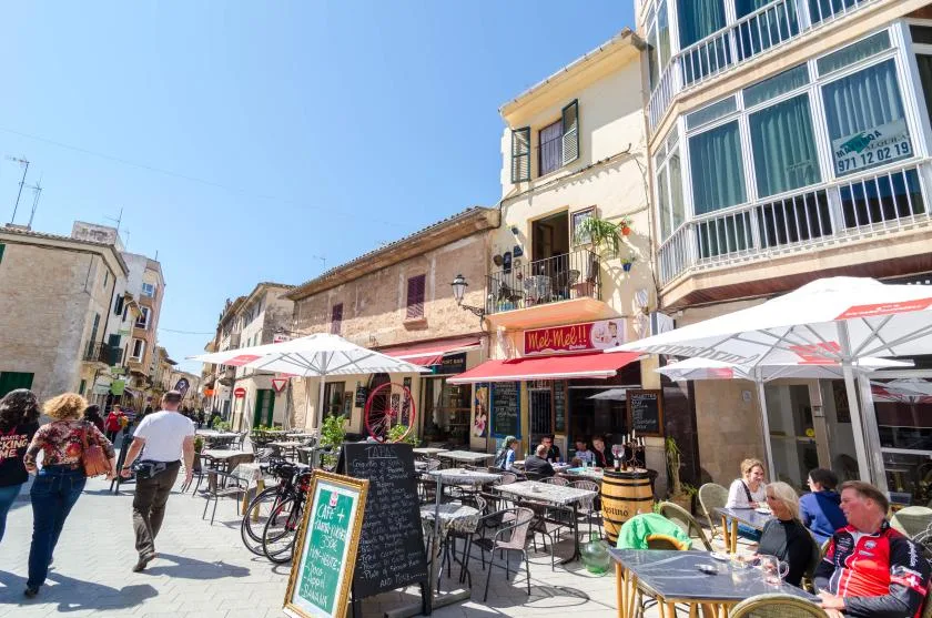 Mesas al aire libre de un café bordean una calle peatonal junto a tiendas y edificios de viviendas.