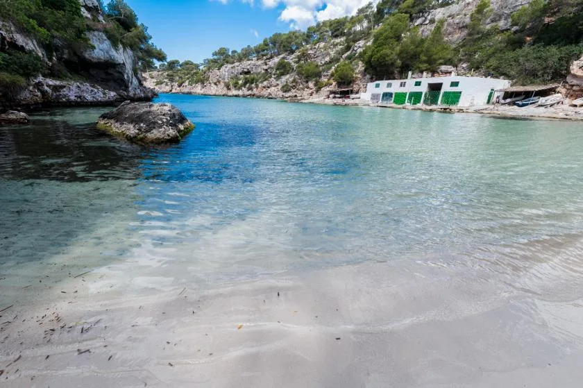 Una cala de aguas claras con orilla arenosa y pequeños edificios junto a la ribera rocosa.