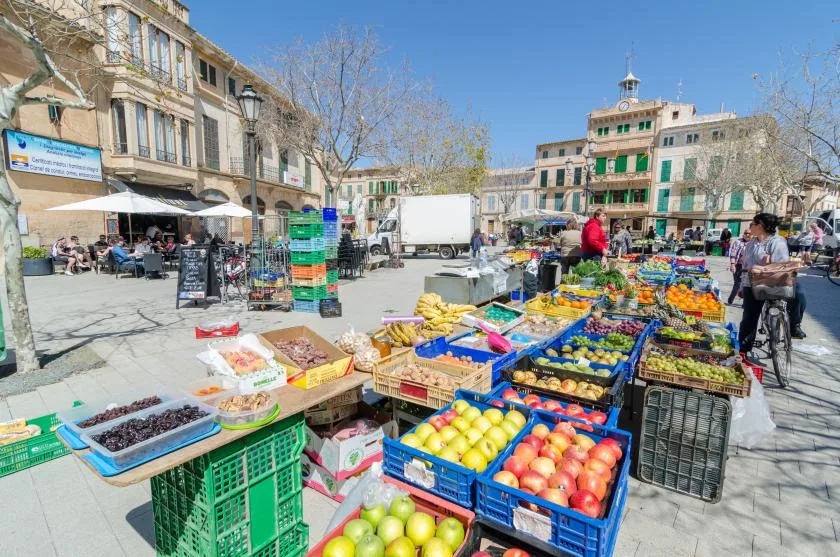 Puestos de mercado al aire libre exhiben frutas y verduras coloridas en una plaza soleada.