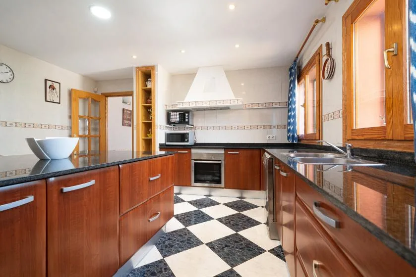 Una cocina luminosa con gabinetes de madera, encimeras oscuras y suelo a cuadros.
