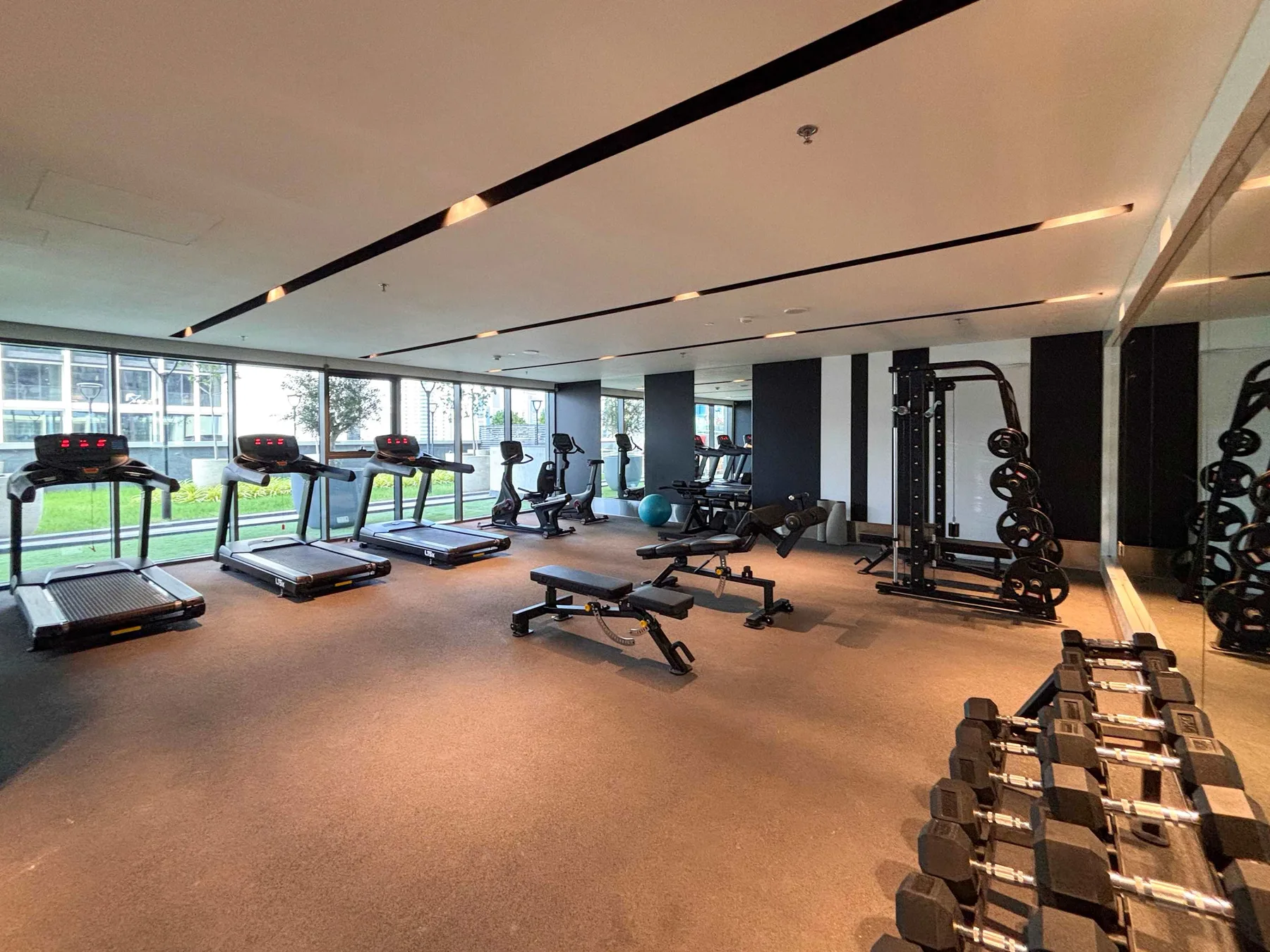 A gym with treadmills, weight benches, and dumbbells beside large windows.