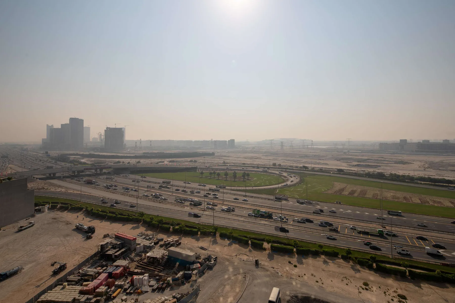 A hazy city skyline overlooks multilane roads and open green land.