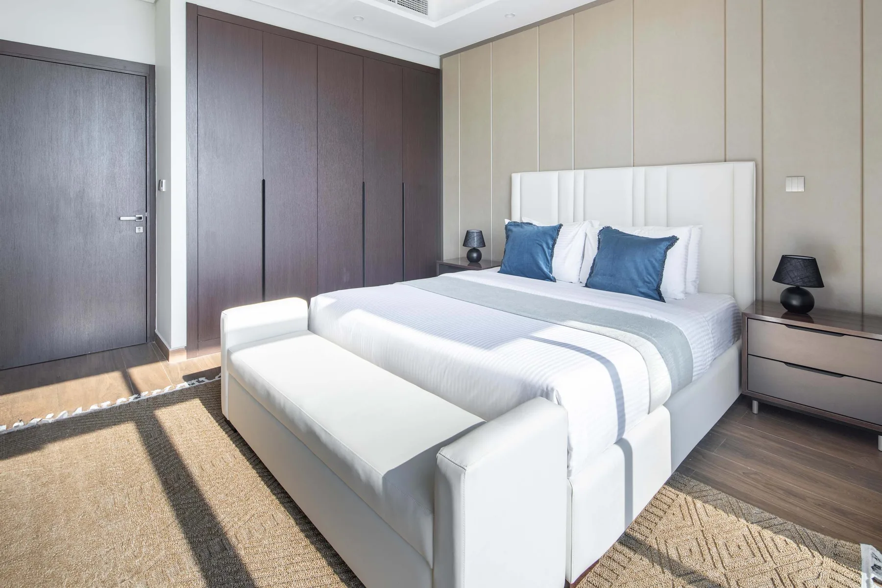 A bright bedroom with a white bed, blue pillows, and dark wardrobes.