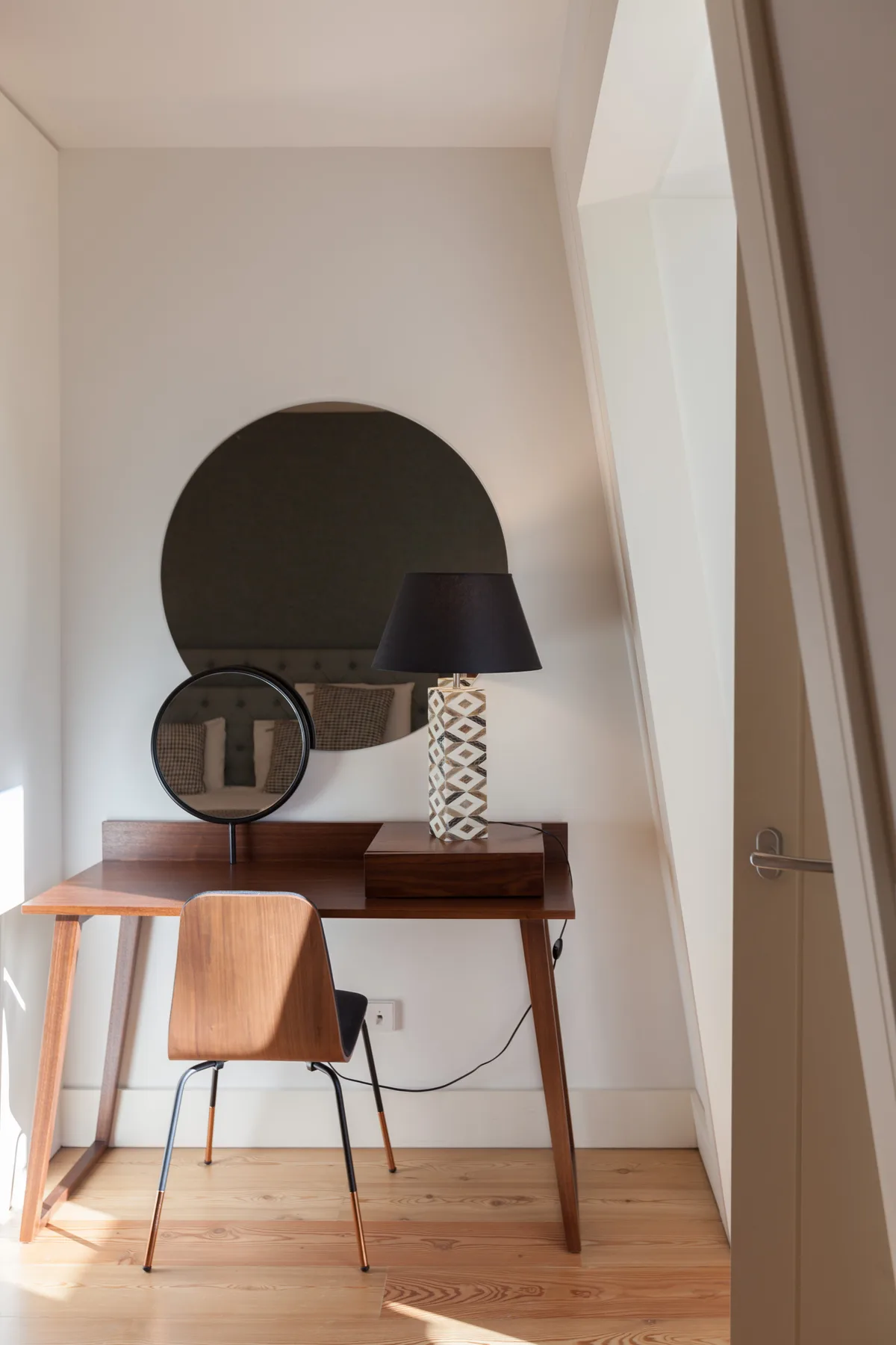 A compact desk area with a chair, lamp, and round mirror.