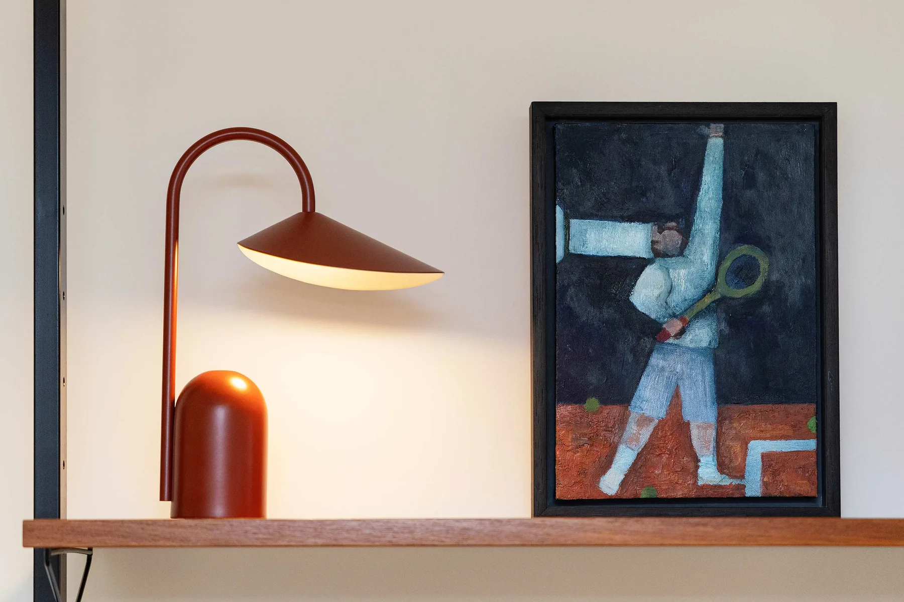 A framed painting and a curved lamp sit on a shelf against a light wall.