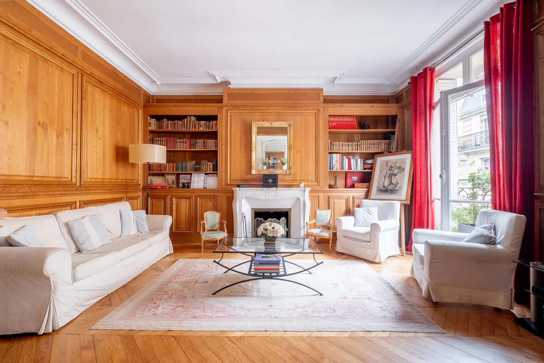 A wood-paneled living room with seating, bookshelves, and tall windows with red curtains.