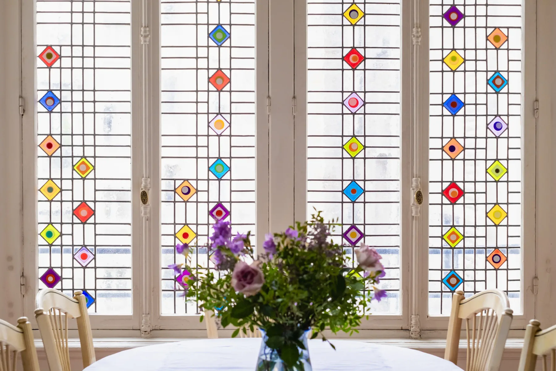A dining table with flowers stands before tall windows decorated with colorful geometric glass.