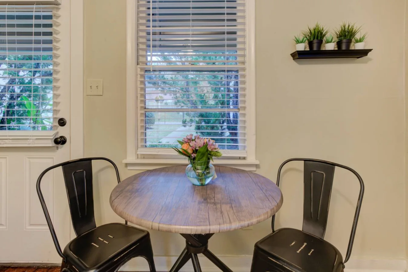 A small dining area has a round wooden table and two chairs beside windows.
