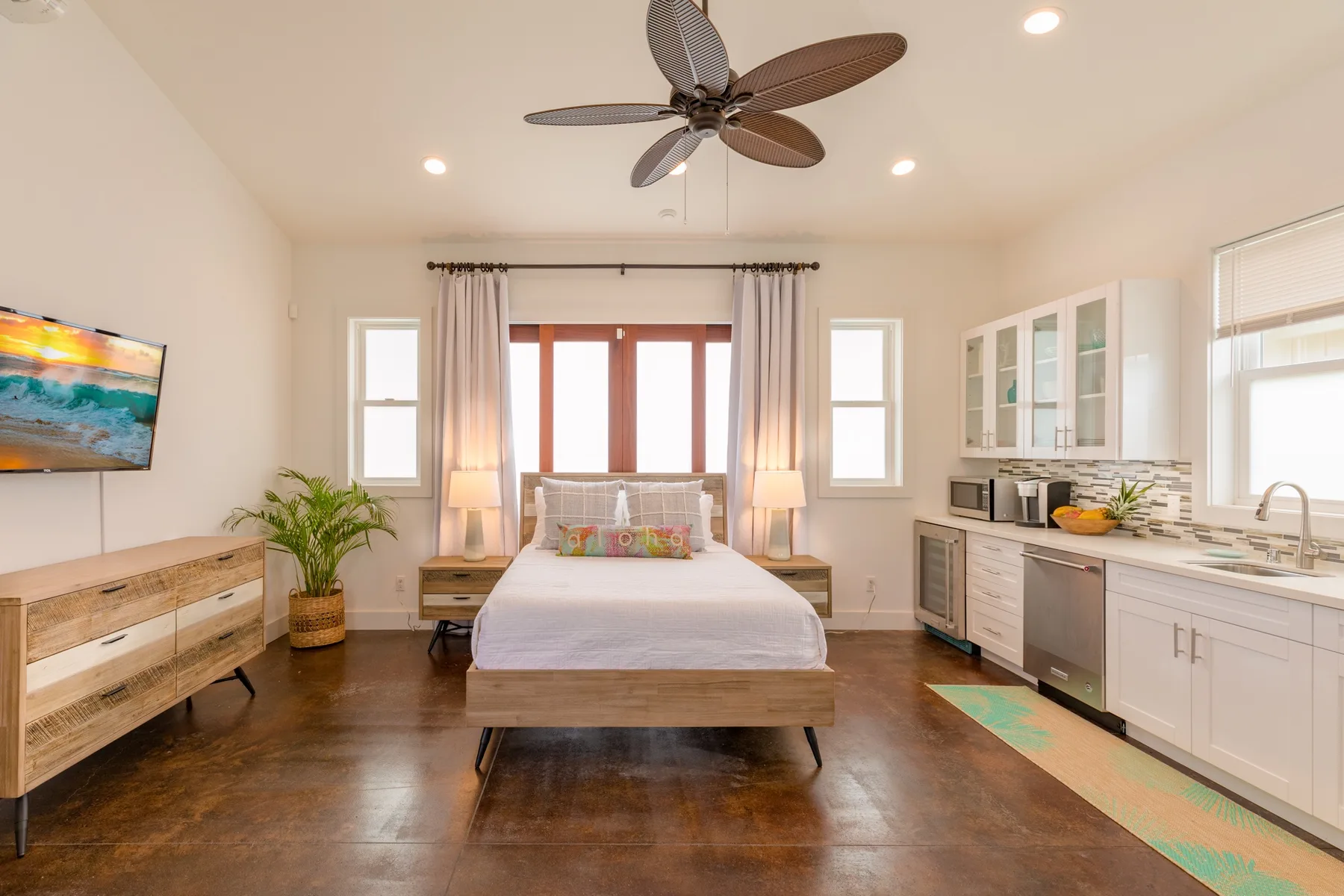 A bright bedroom with a bed, curtained windows, and a small kitchenette.