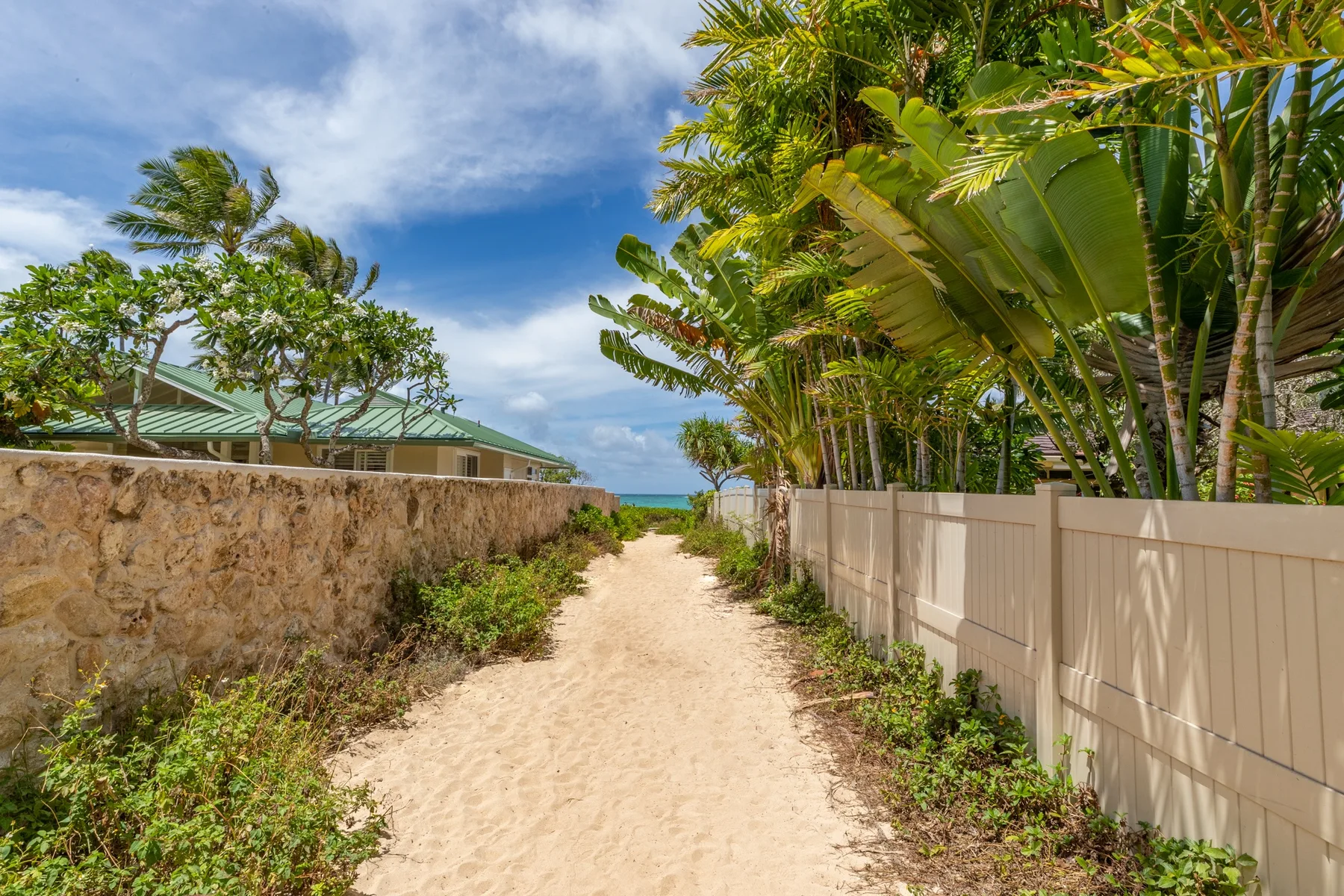 A sandy path runs between a stone wall and a white fence, with tropical greenery.