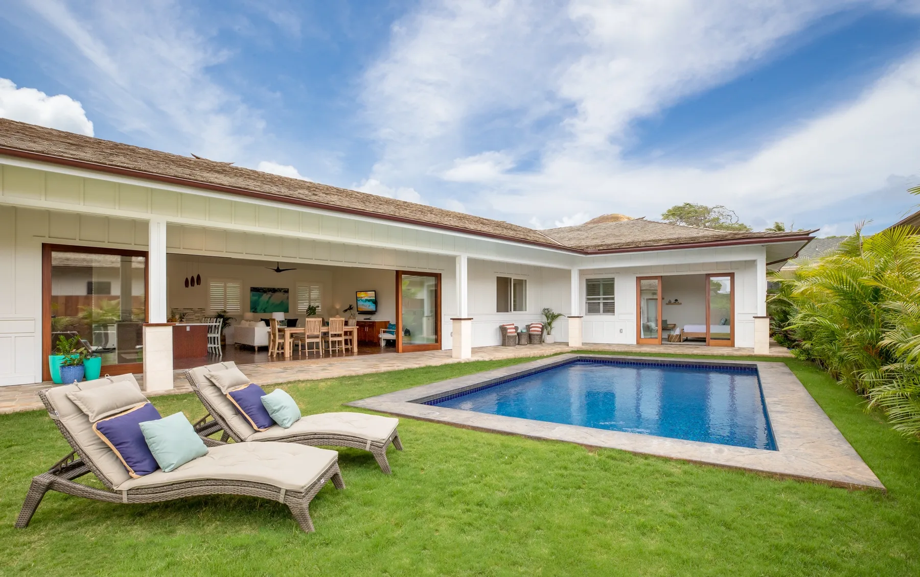 A house opens onto a backyard pool, with outdoor seating on the lawn.