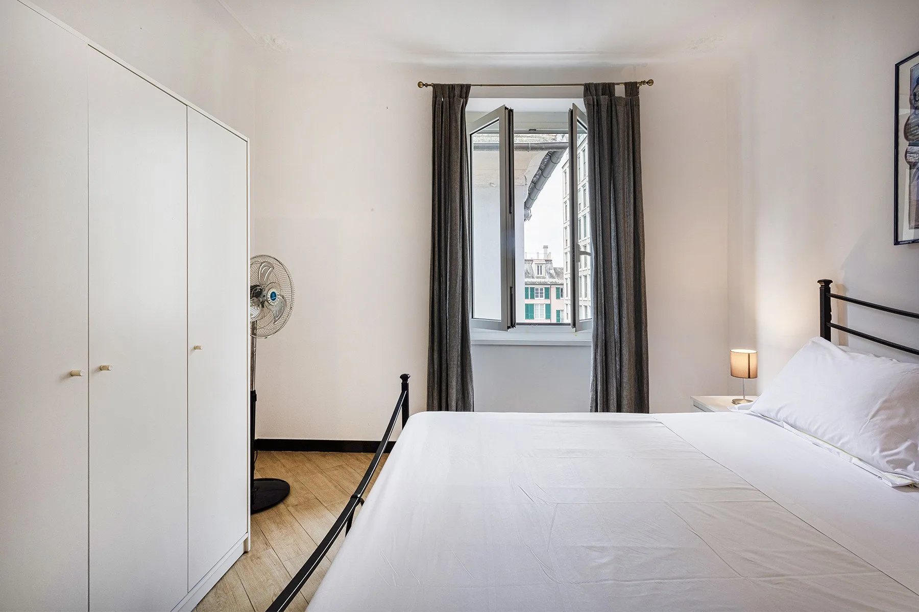 A bright bedroom with a bed, wardrobe, and open window framed by curtains.