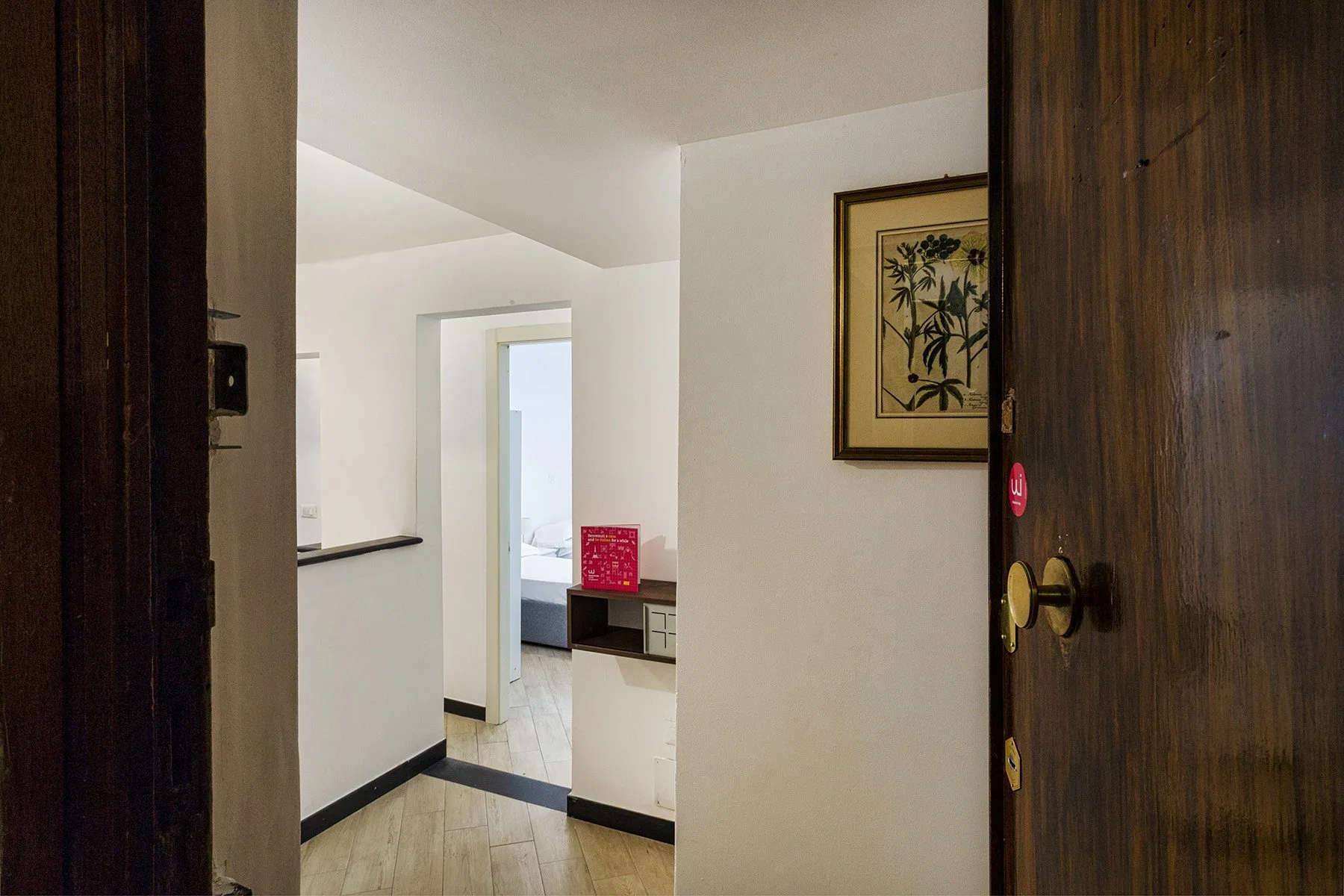 A bright interior hallway opens toward a room with a bed and framed botanical artwork.