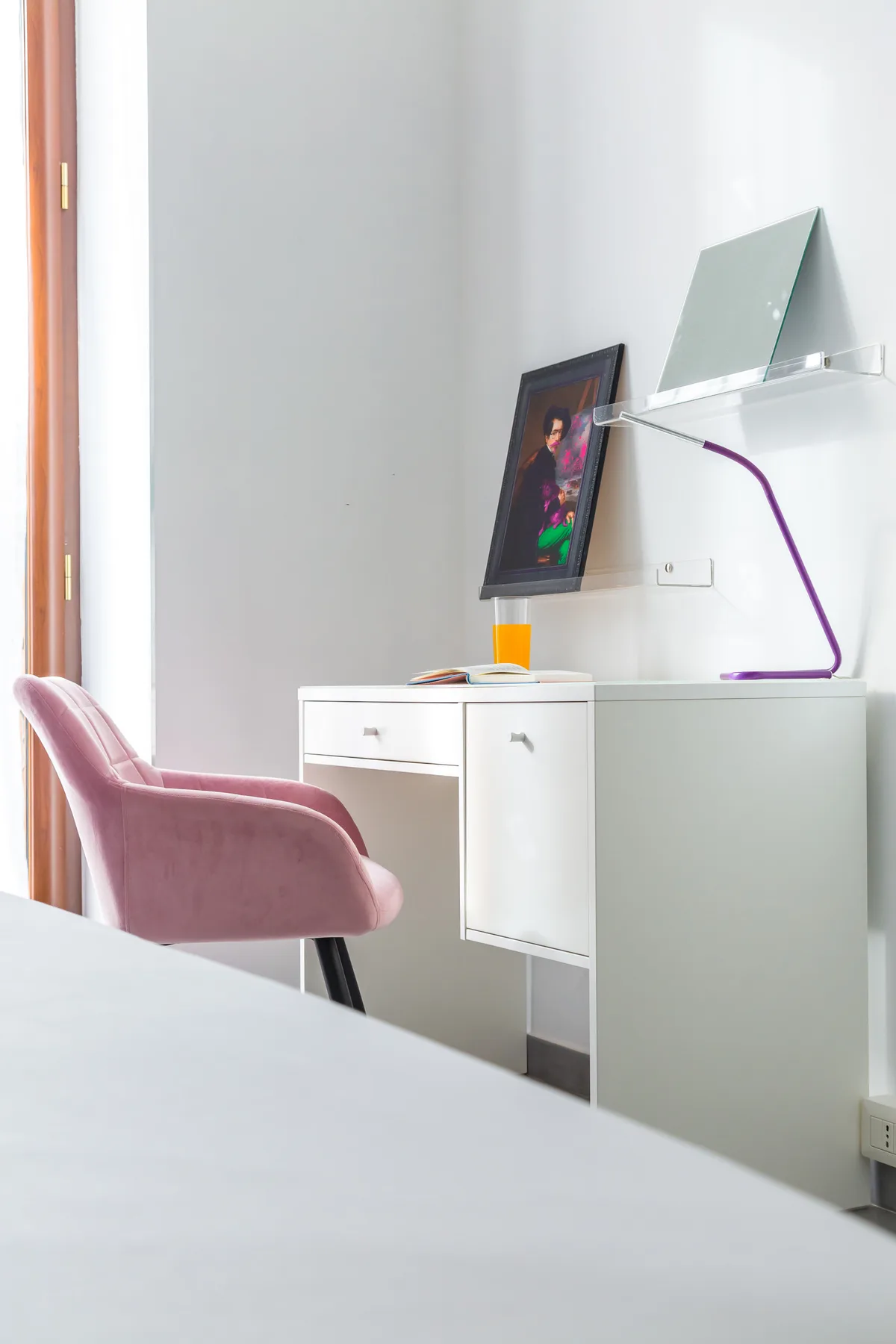 A desk with a pink chair and a framed picture stands beside a bright window.