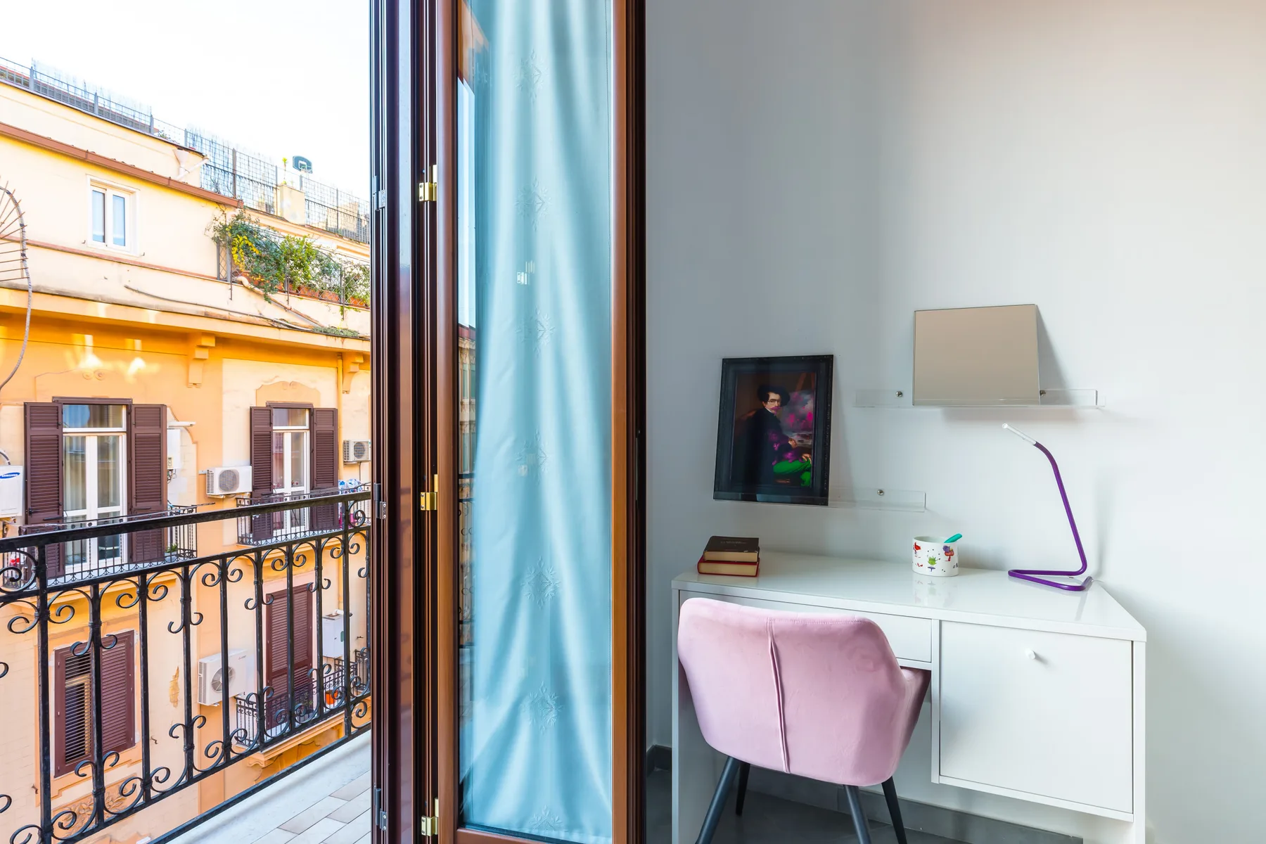 A desk and seating sit beside a glass balcony door overlooking neighboring buildings.