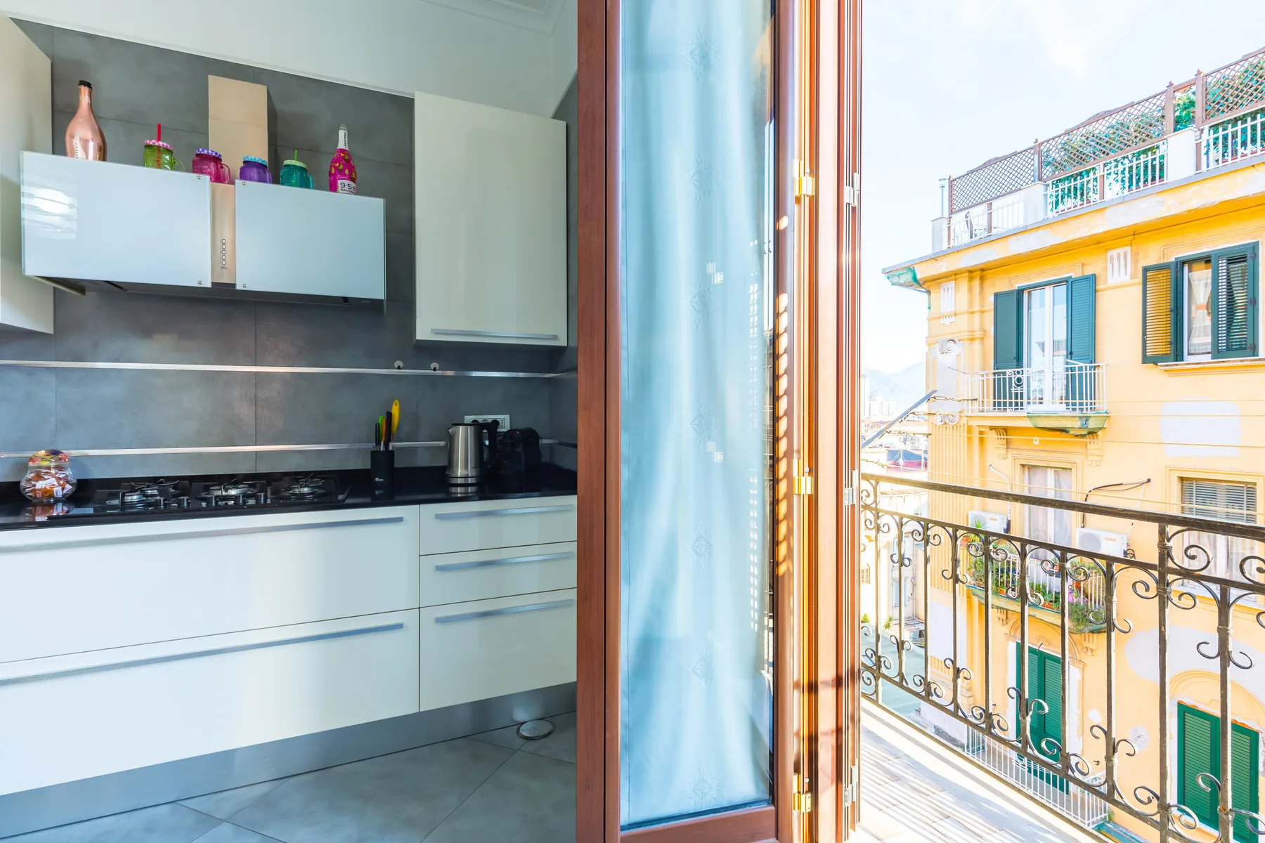 A kitchen with white cabinets opens onto a balcony overlooking nearby buildings.