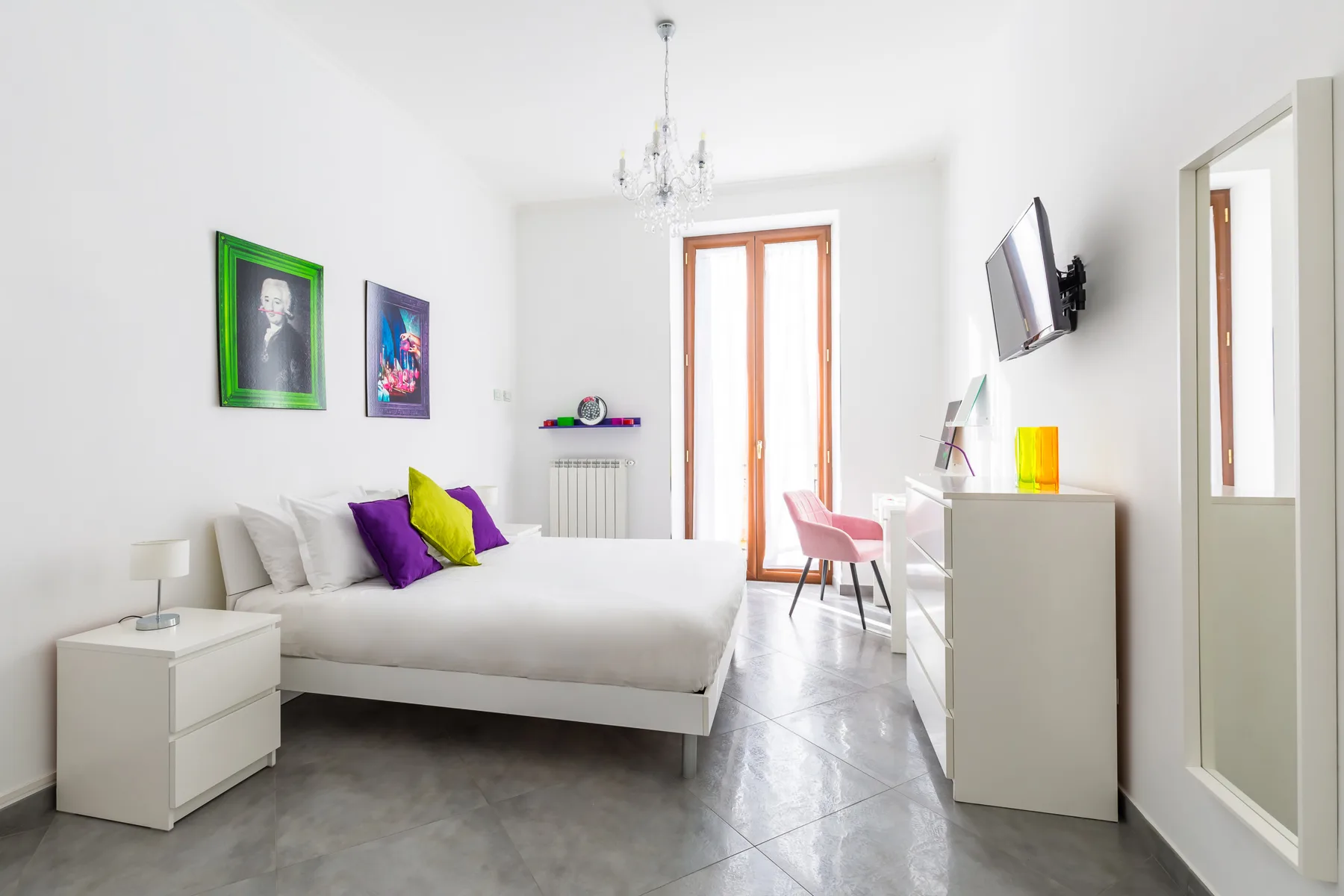 A bright bedroom with a white bed, colorful wall art, and a tall window.
