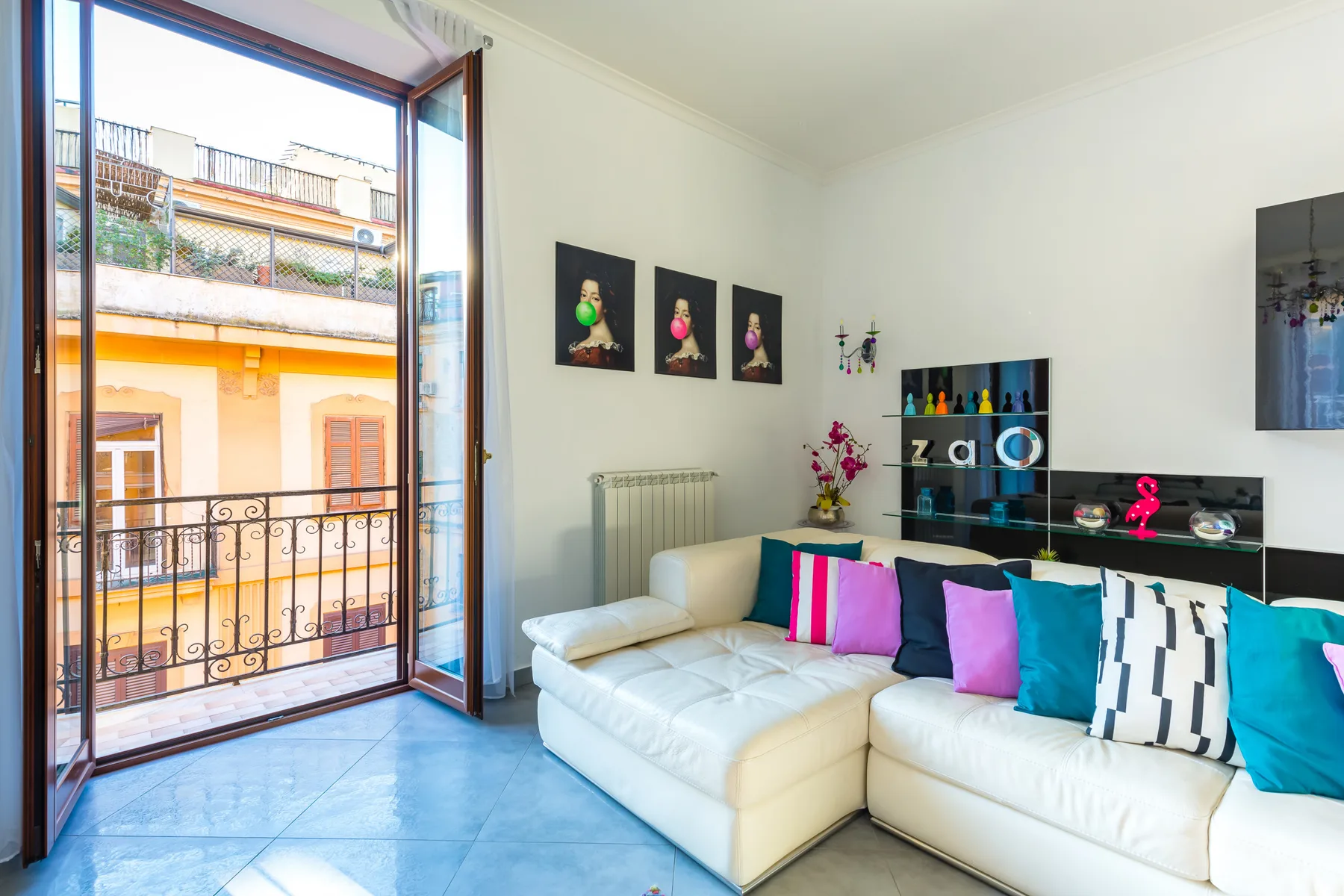 A bright living room with white seating, colorful cushions, and an open balcony door.